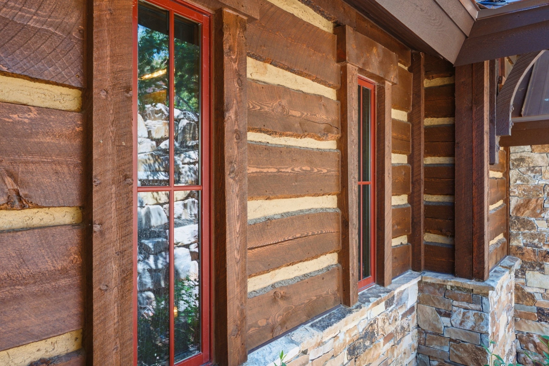 Rustic log cabin exterior featuring chinked log construction, red-framed multi-pane windows, natural stone wainscoting, and wood trim detailing