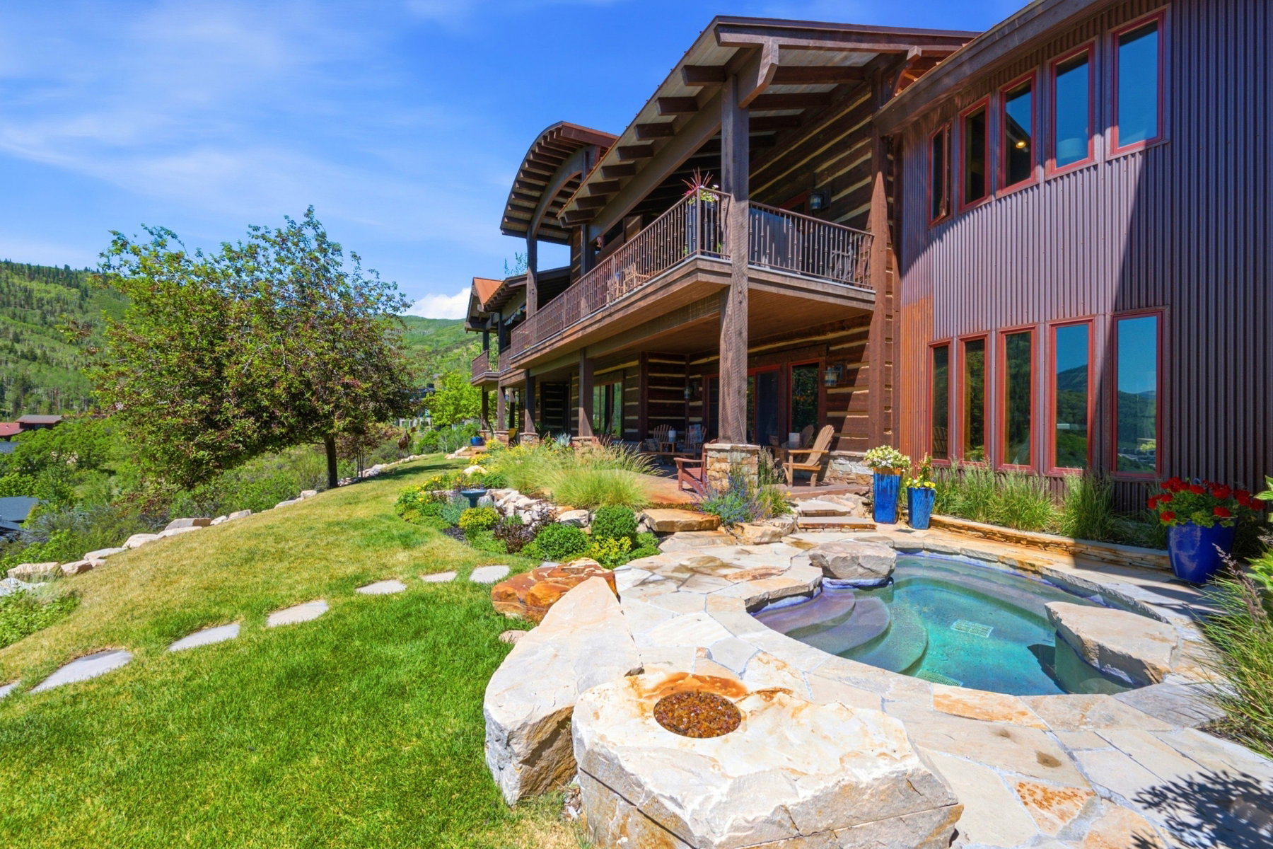 Multi-level exterior featuring a stone-surrounded spa with integrated fire pit, expansive lawn, and mature landscaping