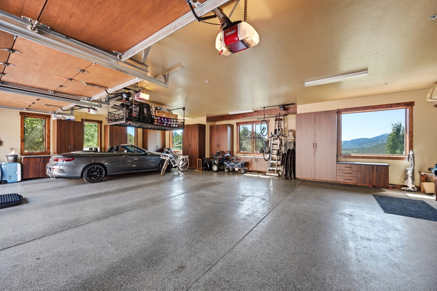 Spacious garage featuring a durable epoxy floor, extensive built-in cabinetry, multiple windows with natural light, overhead storage racks, and fluorescent lighting