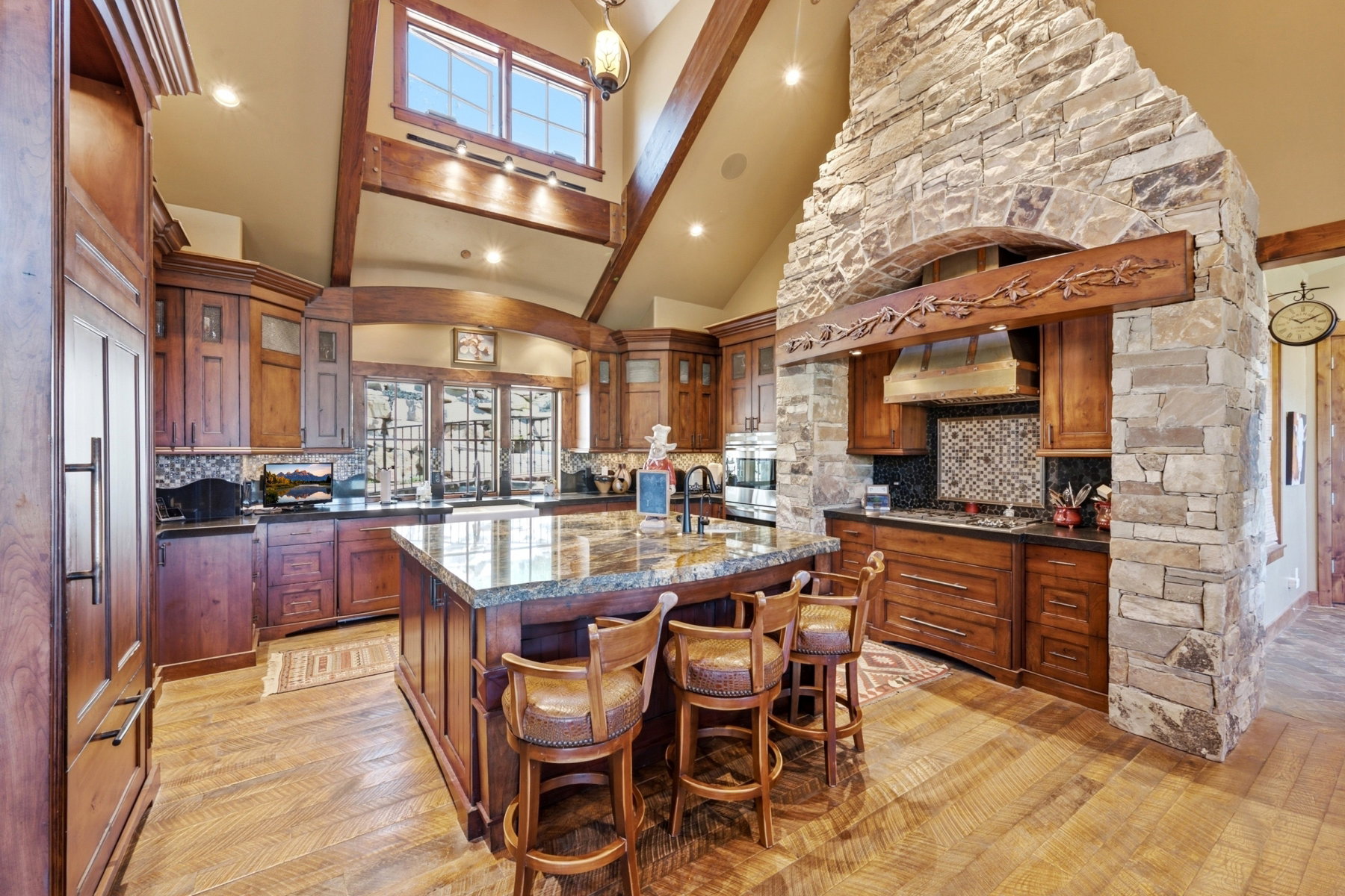 Gourmet kitchen featuring a large central island with a granite countertop, custom wood cabinetry, stainless steel appliances, a stone-clad range hood, and wood-finish flooring