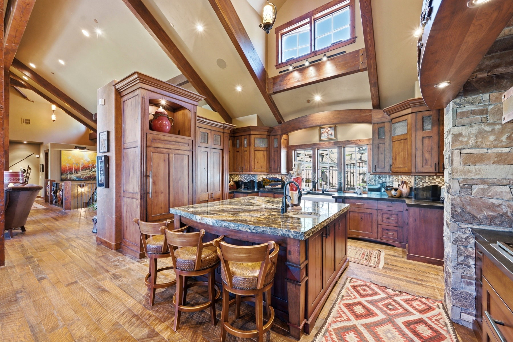 Kitchen featuring a large island with a natural stone countertop, custom wood cabinetry, wood-finish flooring, exposed wood beams, and a natural stone wall accent