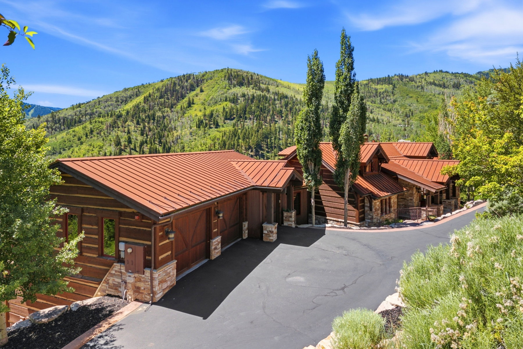 Mountain-style residence featuring log siding, multiple gables with a red metal roof, a paved driveway, and stone accents