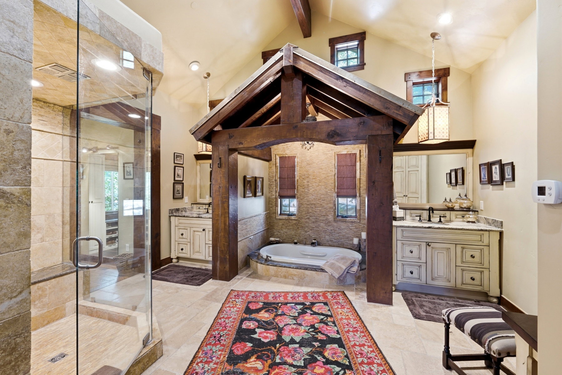 Expansive bathroom featuring a prominent timber-frame structure over the soaking tub, a spacious walk-in shower with stone-tiled walls, dual vanity cabinets with stone countertops, and vaulted ceilings with exposed wood beams