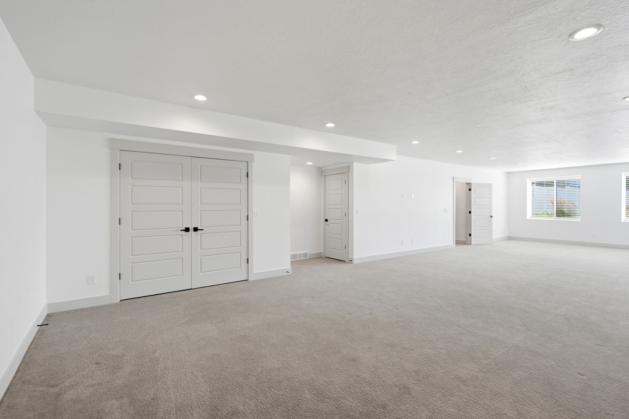 Spacious room featuring light gray carpeting, recessed lighting, white walls, and a pair of double doors with black hardware