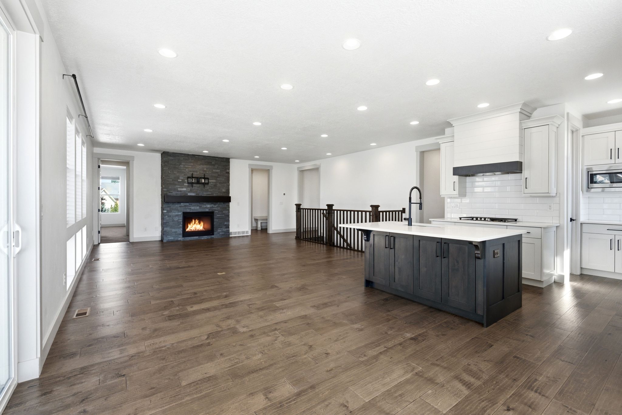 Spacious interior with wood-finish flooring, a contemporary kitchen island with a dark finish, white perimeter cabinetry, a subway tile backsplash, and a stone-clad fireplace