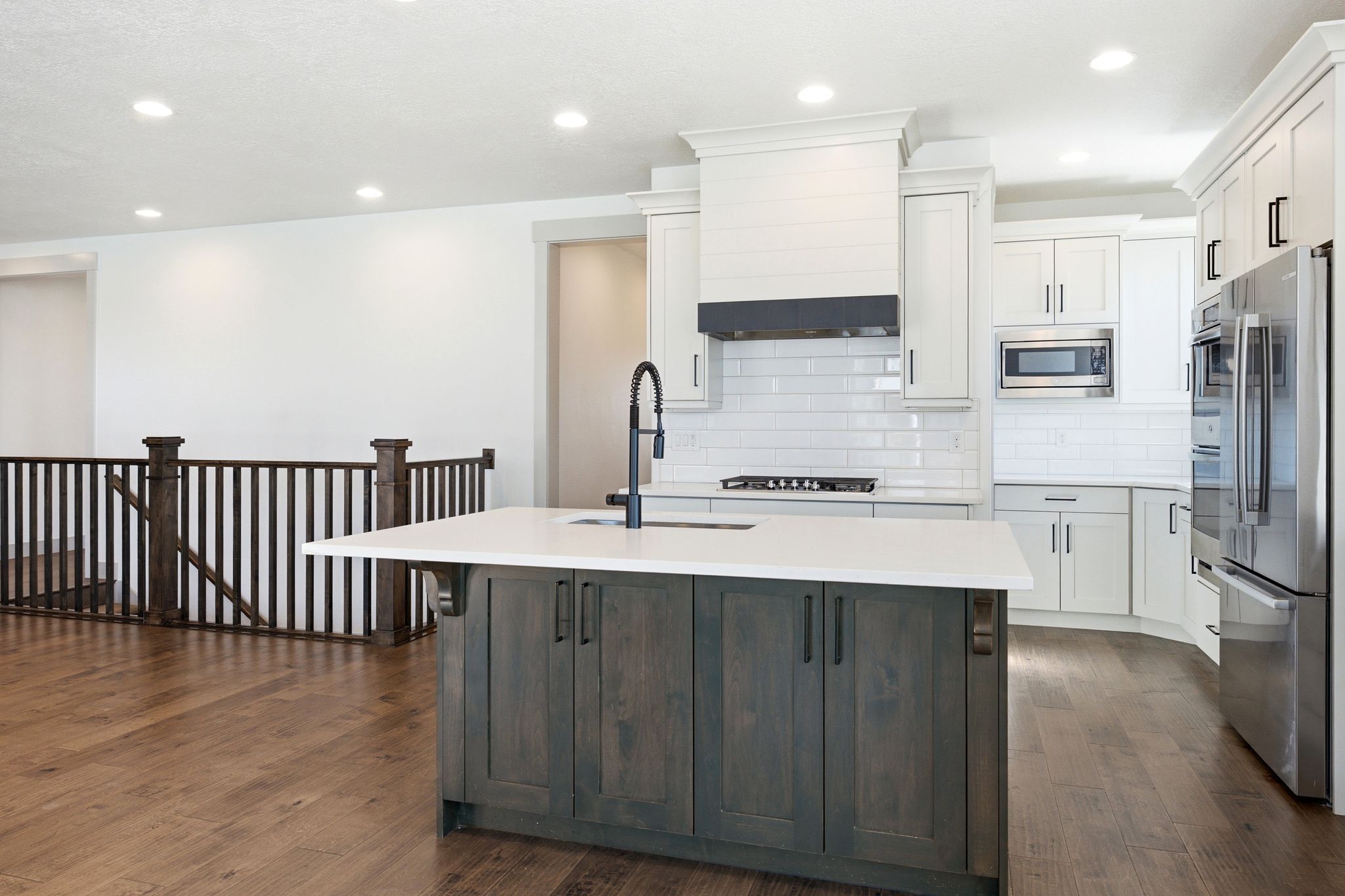 Kitchen featuring a central island with a sink and dark wood-finish cabinetry, light stone-finish countertops, white subway tile backsplash, and stainless steel appliances including a refrigerator, double oven, and microwave