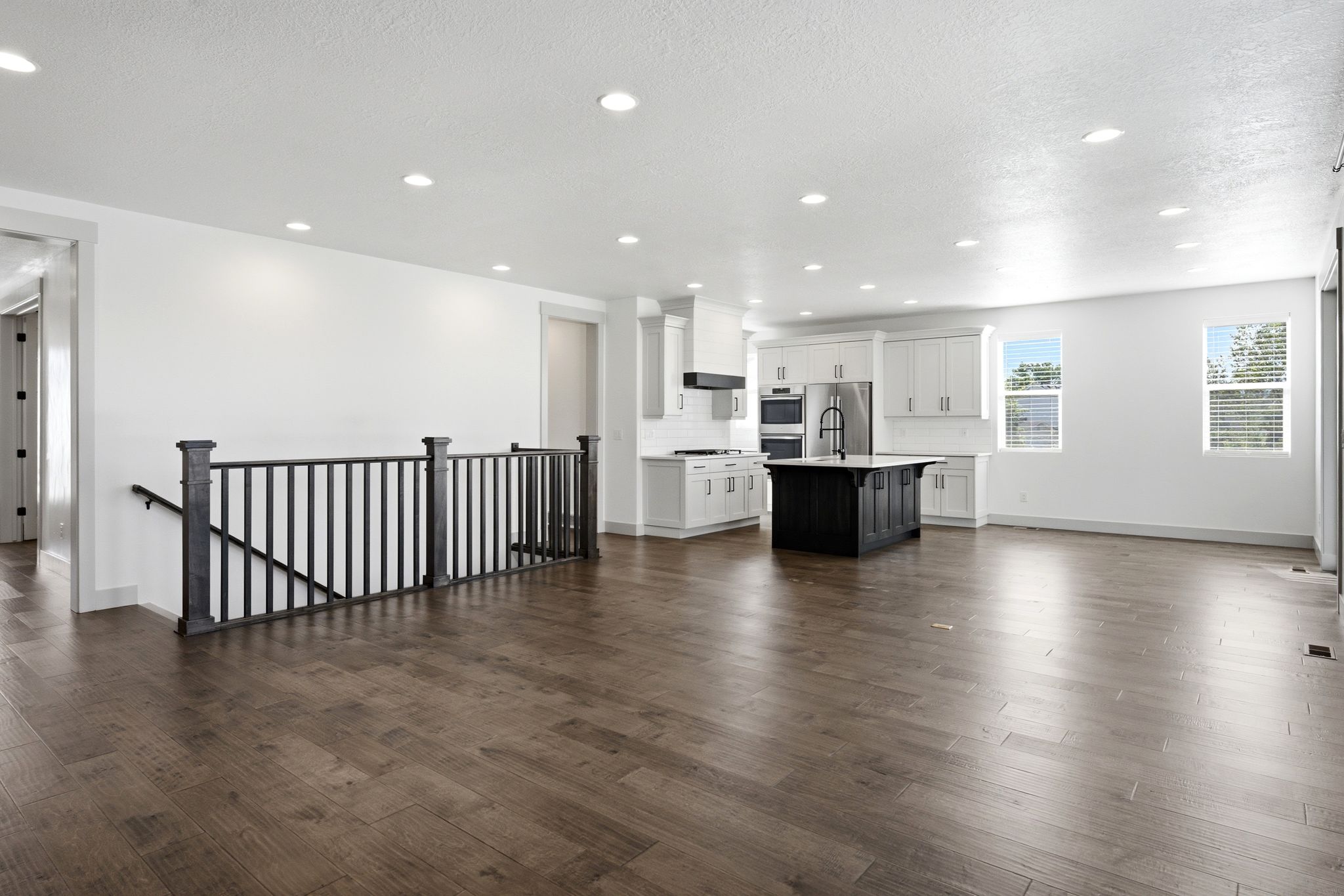 Expansive open-concept living space featuring wood-finish flooring, a modern kitchen with white cabinetry and a dark-toned island, stainless steel appliances, and recessed lighting