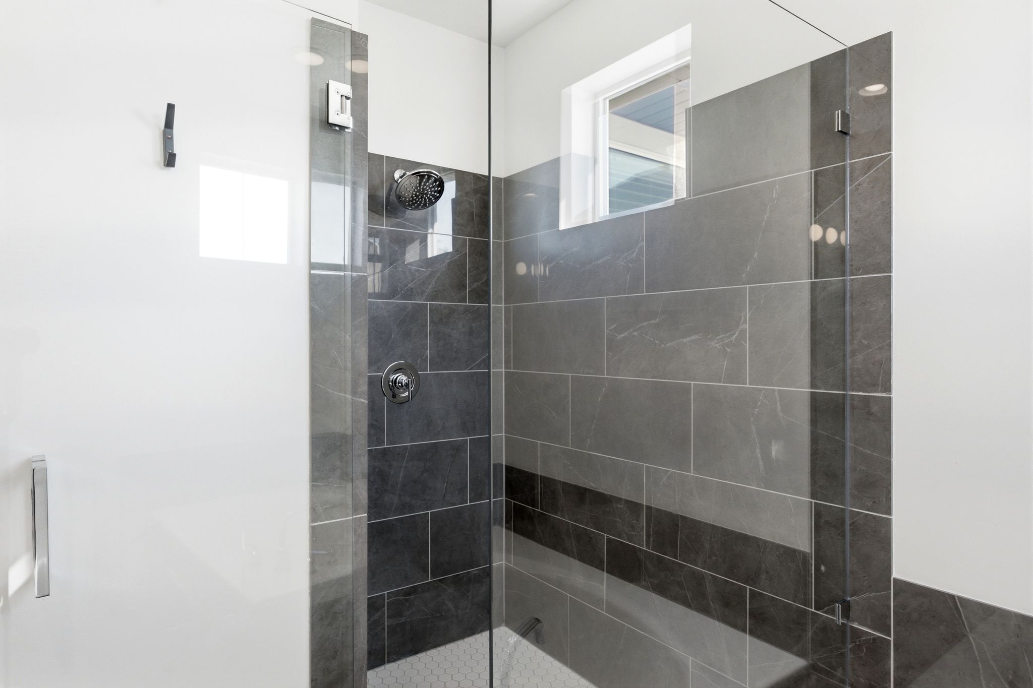 Contemporary glass-enclosed shower featuring large format gray tile, a contrasting dark gray accent tile band, chrome shower fixtures, and a high-set window