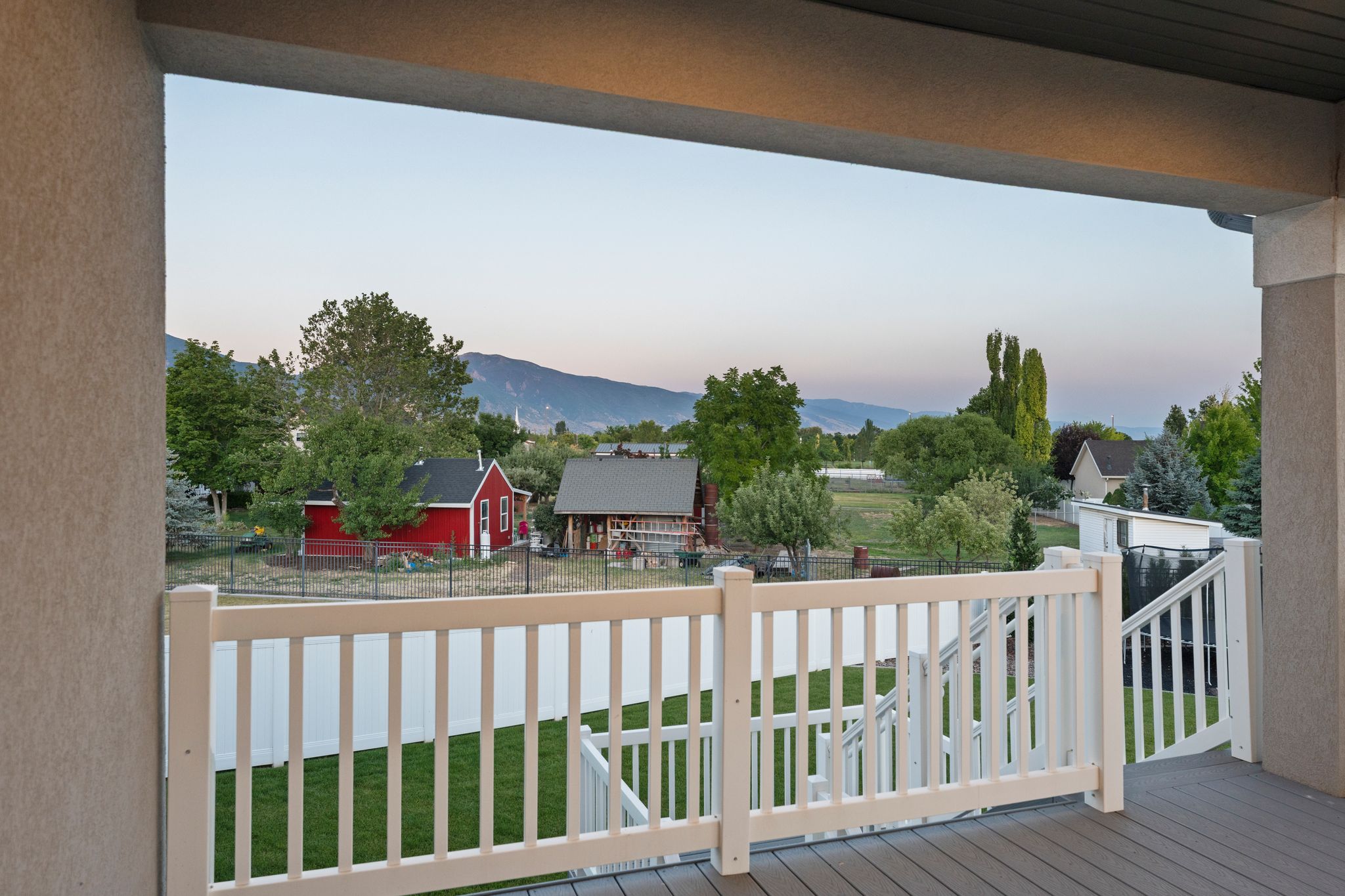 Covered porch with composite decking and white railing, overlooking a green lawn, distant red outbuilding, and tree-lined landscape with mountains