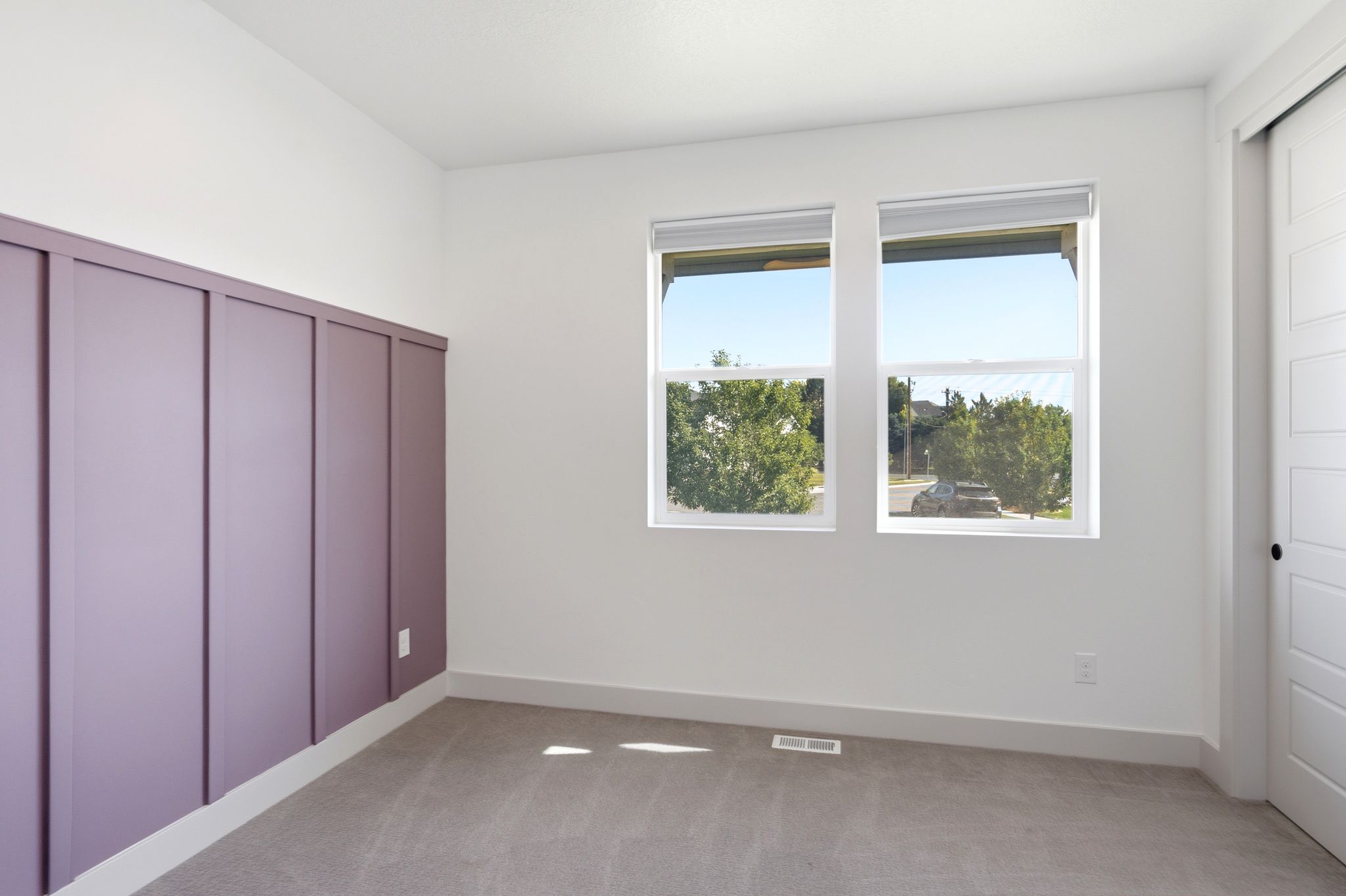 Light-filled interior featuring a lavender accent wall with vertical paneling, two white-framed windows, and gray carpet flooring