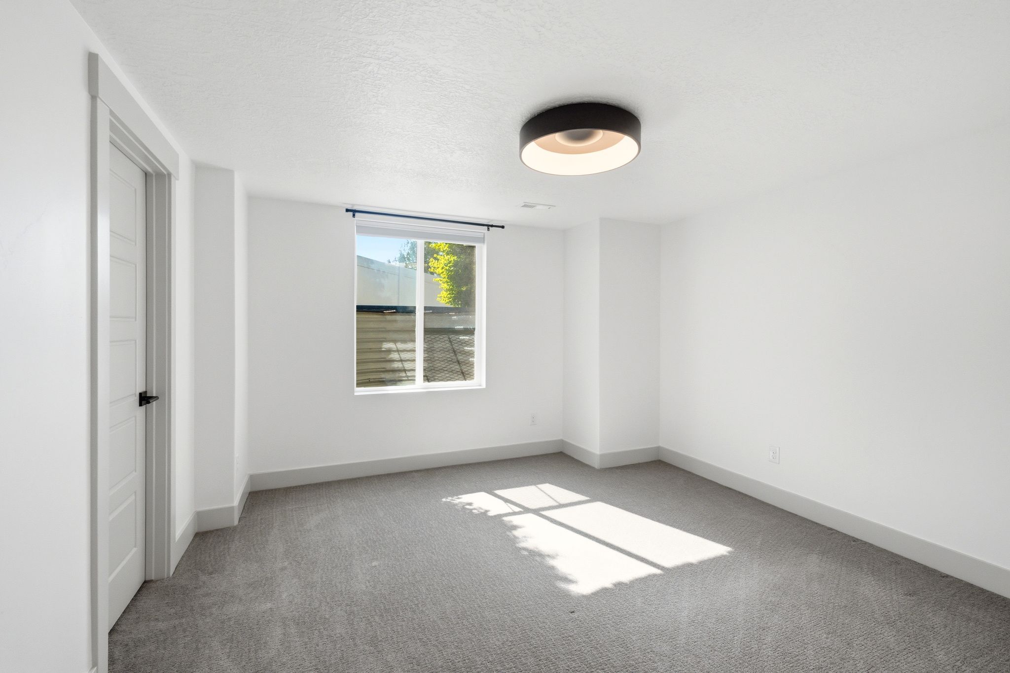 Room featuring a single window with roller shade, gray carpet, white walls, and a modern flush-mount ceiling light