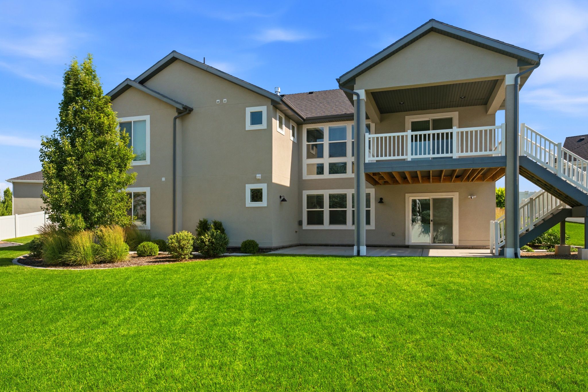 Expansive green lawn bordering the rear of the property, featuring a covered upper deck with white railing, an exterior staircase, and a lower-level patio with sliding glass door access