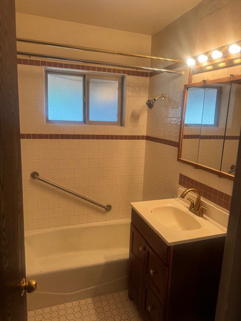 Bathroom featuring a full tub with shower, white subway tile surround with accent banding, and a white vanity with a brass-finish faucet