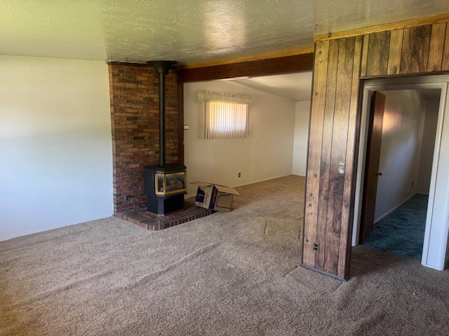 Interior space featuring a brick fireplace with a black wood stove, carpet flooring, a wood-paneled wall, a wood beam, and a window with horizontal blinds