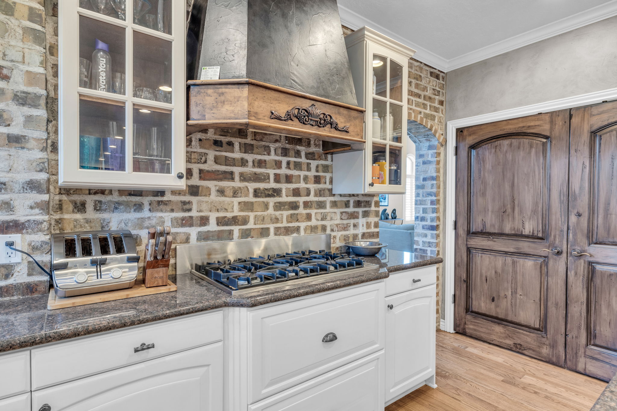 Kitchen featuring a gas cooktop with a decorative wooden hood, brick backsplash, white cabinetry with glass-front uppers, granite countertops, and wood-finish flooring