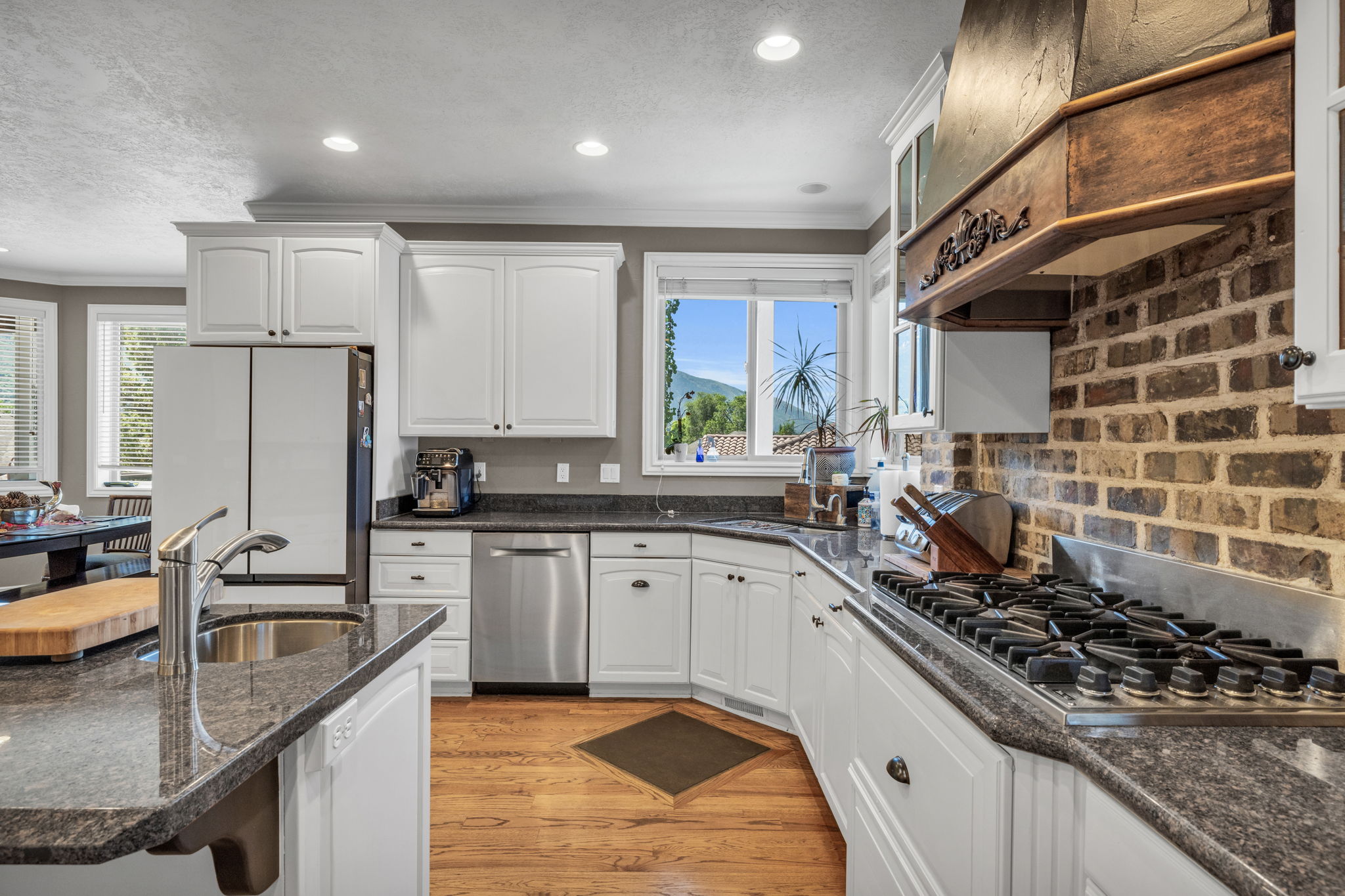 Kitchen featuring white cabinetry, dark granite-finish countertops, stainless steel appliances, wood-finish flooring, and a brick-finish backsplash with a custom wood range hood