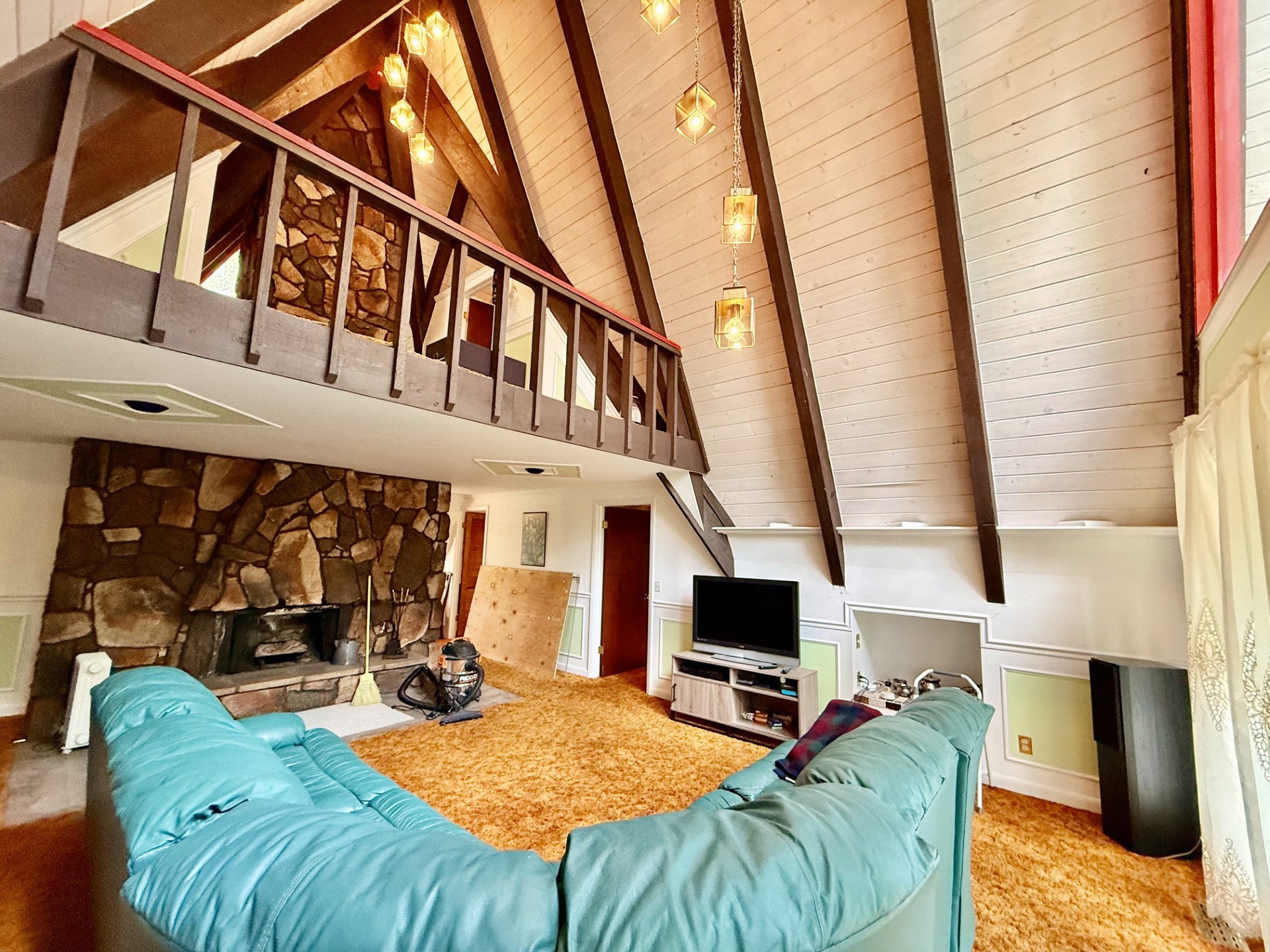 Spacious living area featuring a prominent stone fireplace, a vaulted ceiling with exposed wood beams, and a wood-paneled balcony