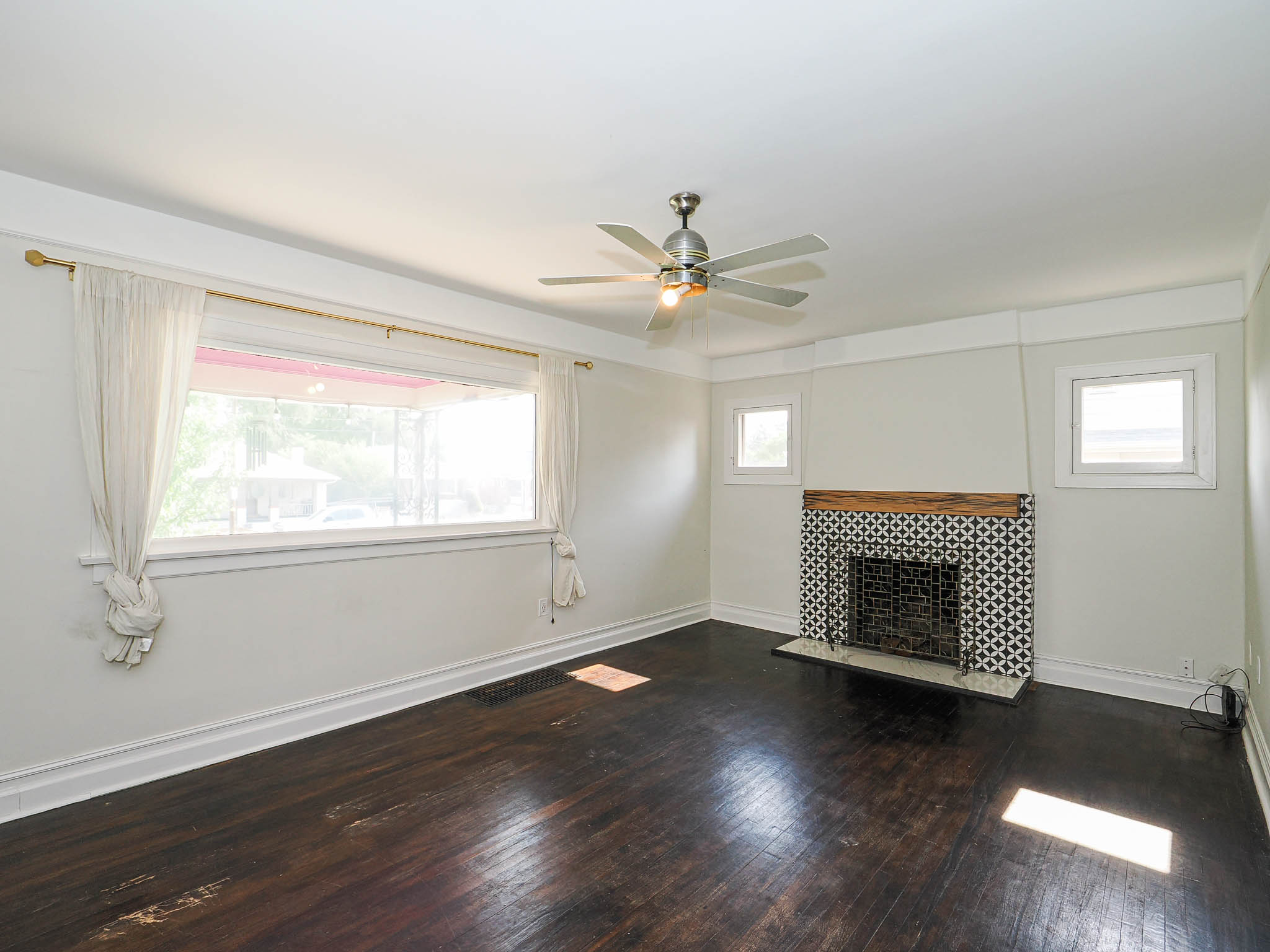 Spacious room featuring dark wood-finish flooring, a decorative fireplace with patterned tile surround and a wood mantel, and a ceiling fan with integrated lighting