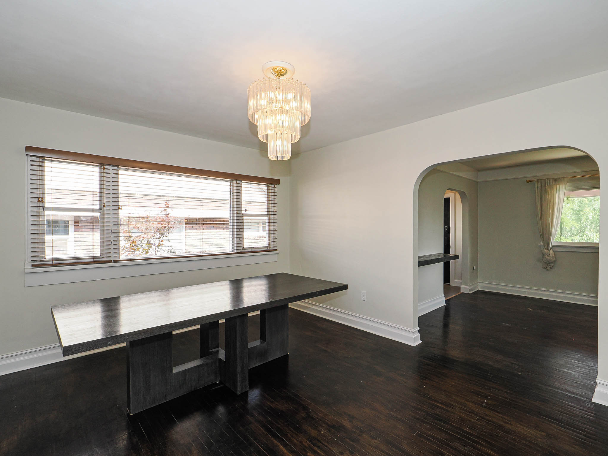 Spacious room featuring dark wood-finish flooring, a tiered chandelier, and a wide window with blinds