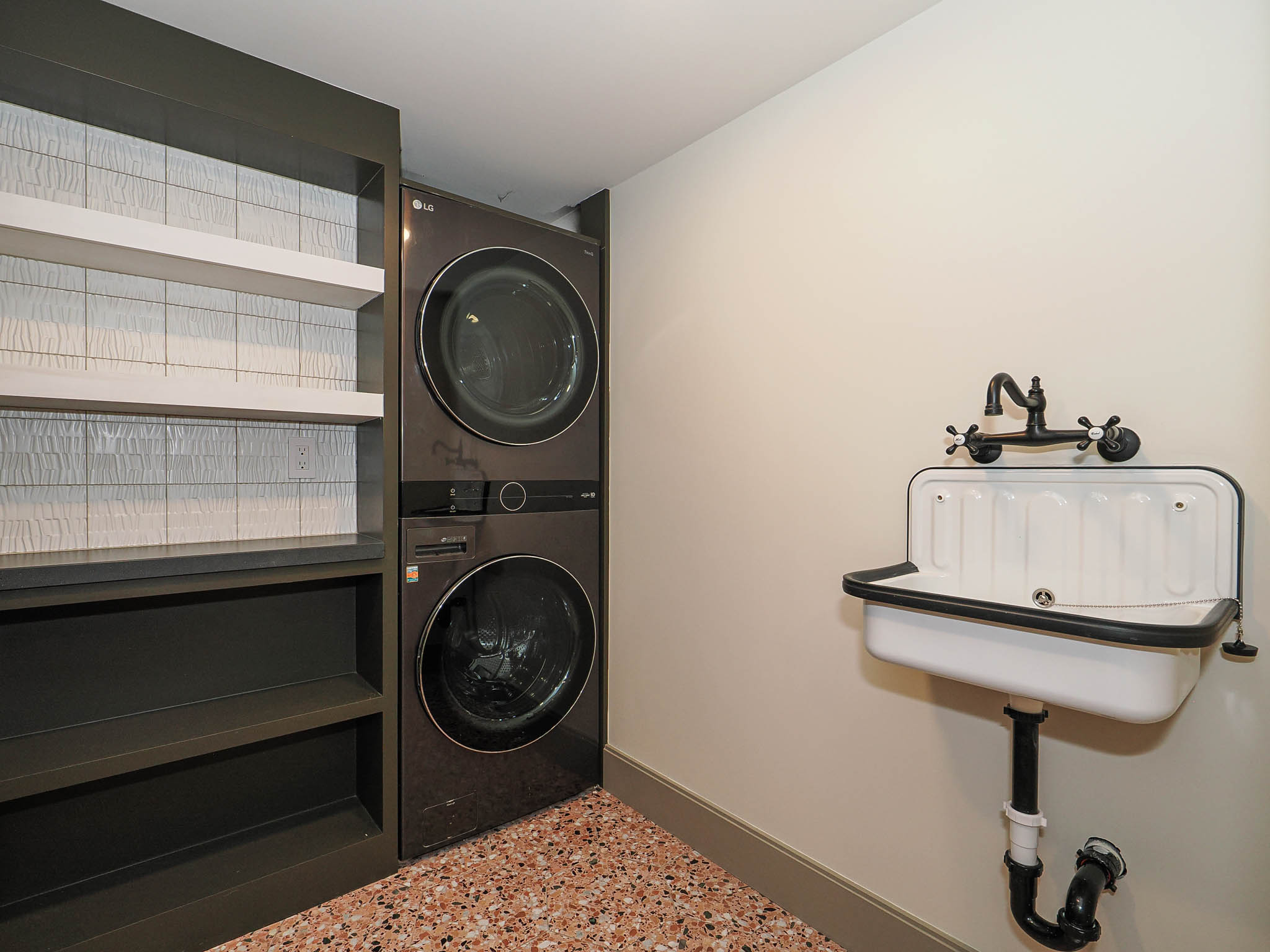 Laundry area featuring a stackable washer and dryer, built-in shelving with textured tile backing, a wall-mounted utility sink with a matte black faucet, and speckled terrazzo flooring