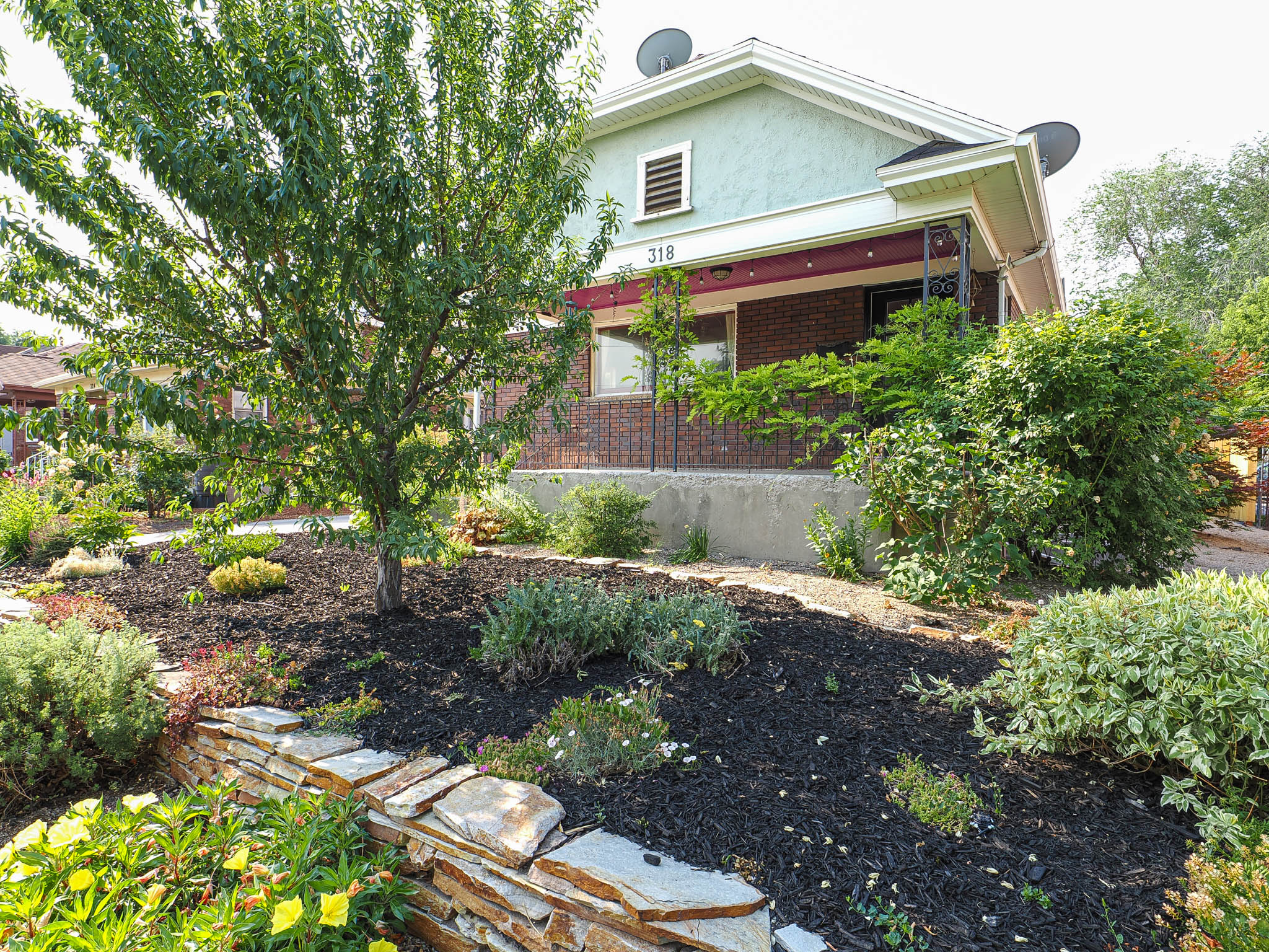 Front landscaping featuring a mature tree, layered plantings, decorative rock wall, and mulched beds