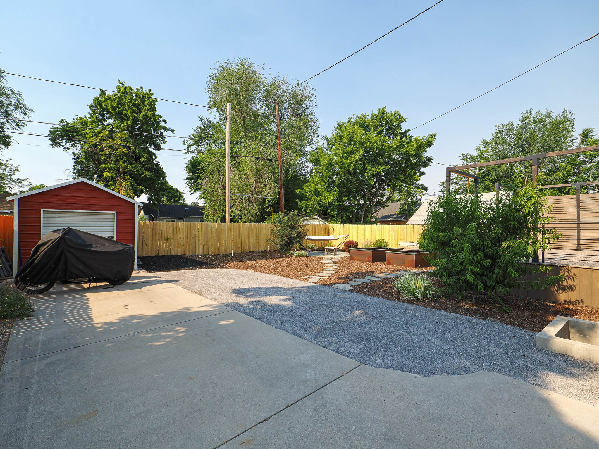 Expansive backyard featuring a concrete patio, gravel pathways, a red storage shed with a roll-up door, a wooden privacy fence, and raised garden beds