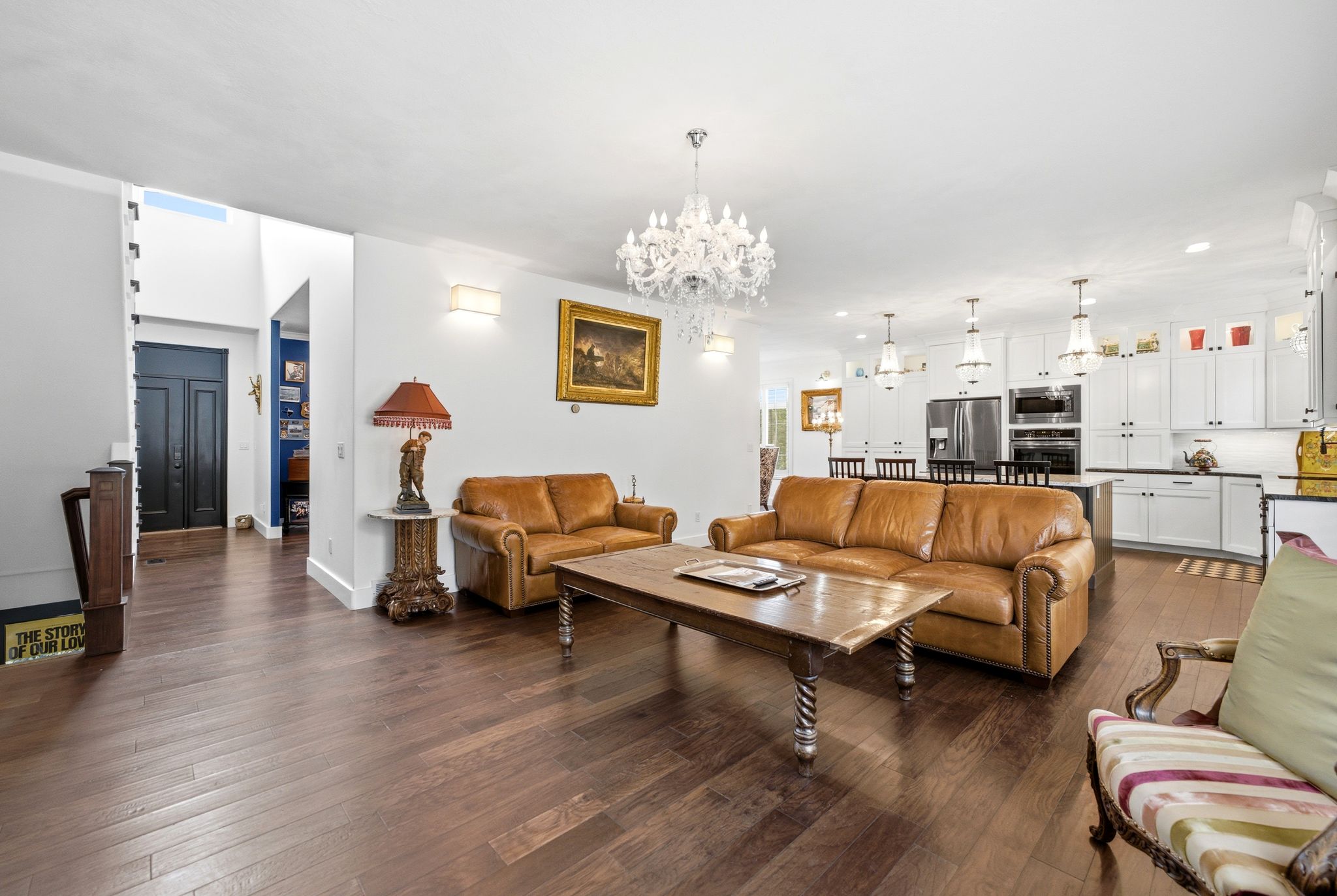 Open-concept living area featuring wood-finish flooring, a white kitchen with stainless steel appliances, a kitchen island with seating, a chandelier, and wall sconces