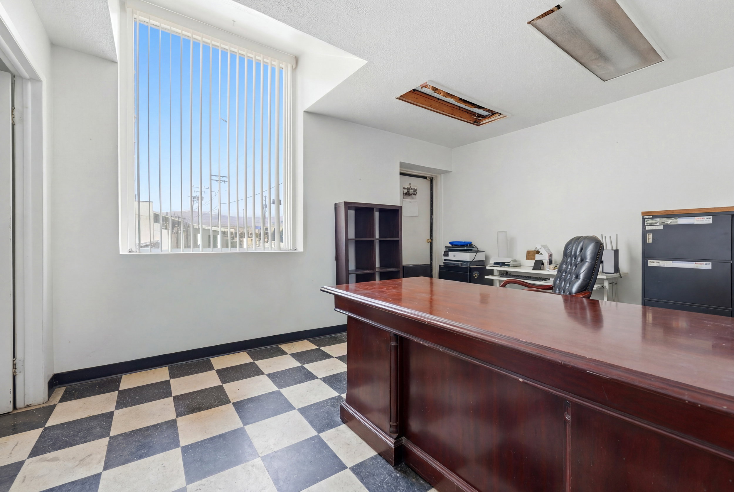 Spacious room featuring a large window with vertical blinds, checkerboard floor tiling, and a built-in wood-finish desk