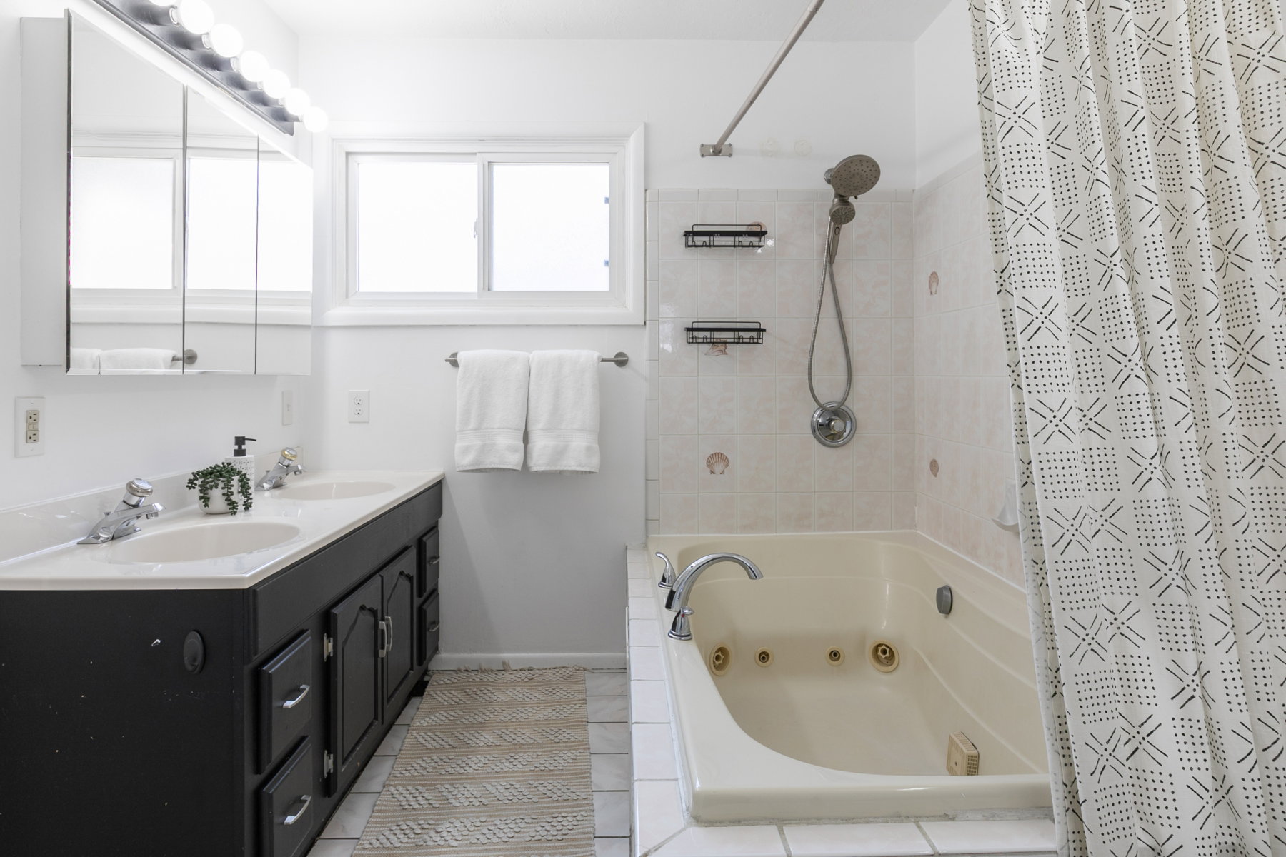 Bathroom featuring a dual sink vanity with a black base cabinet and white countertop, a jetted bathtub, and a tiled shower surround