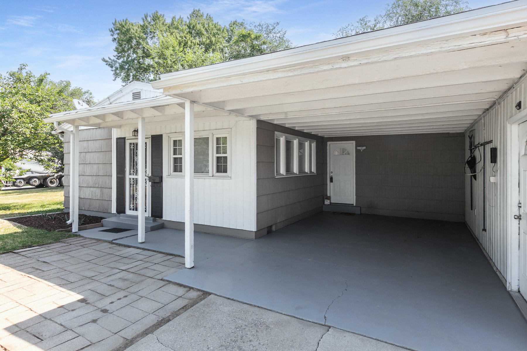 Exterior featuring a covered carport, white trim, gray siding, and a paver stone driveway