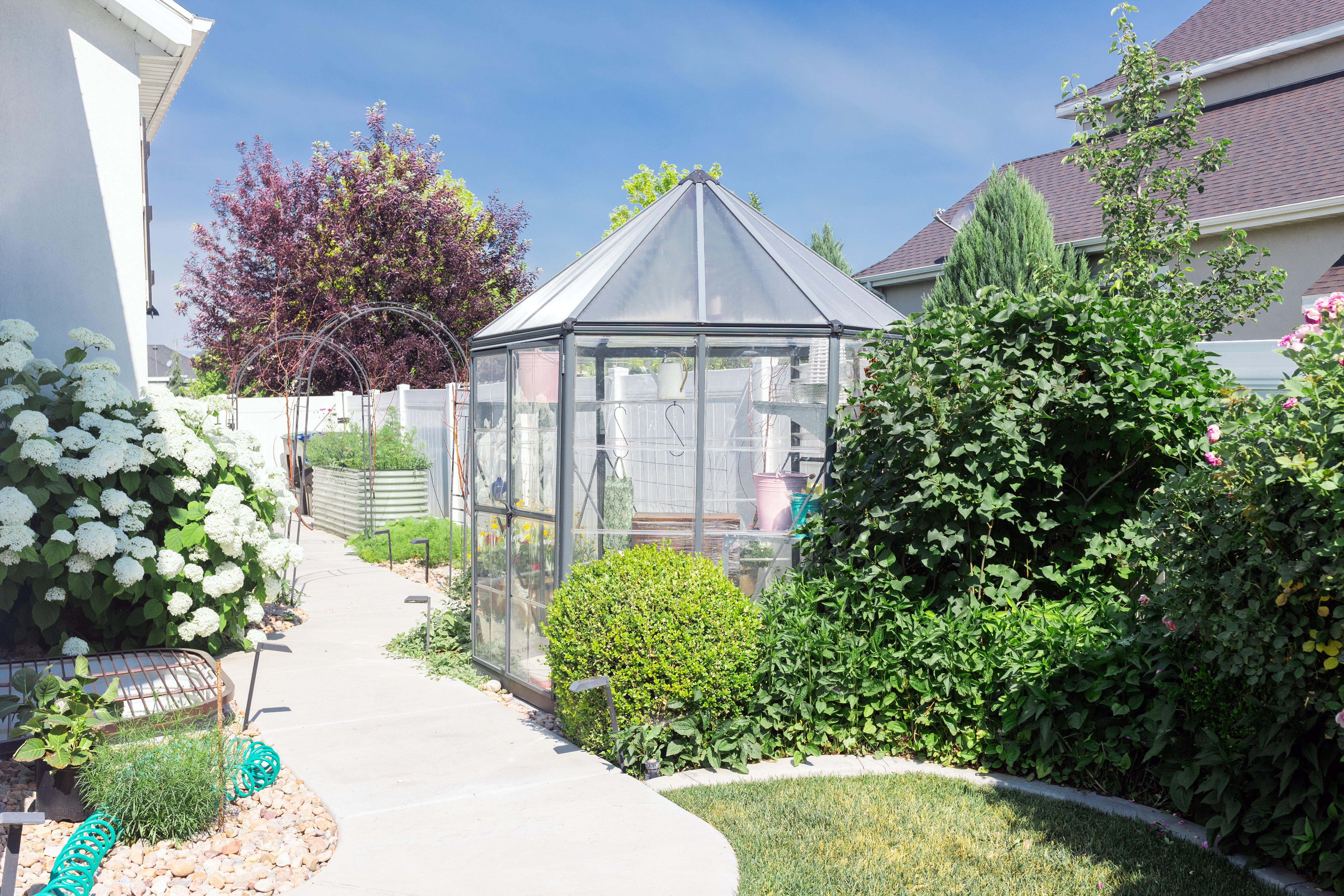Garden featuring a glass greenhouse, concrete pathway, raised garden beds, and mature landscaping