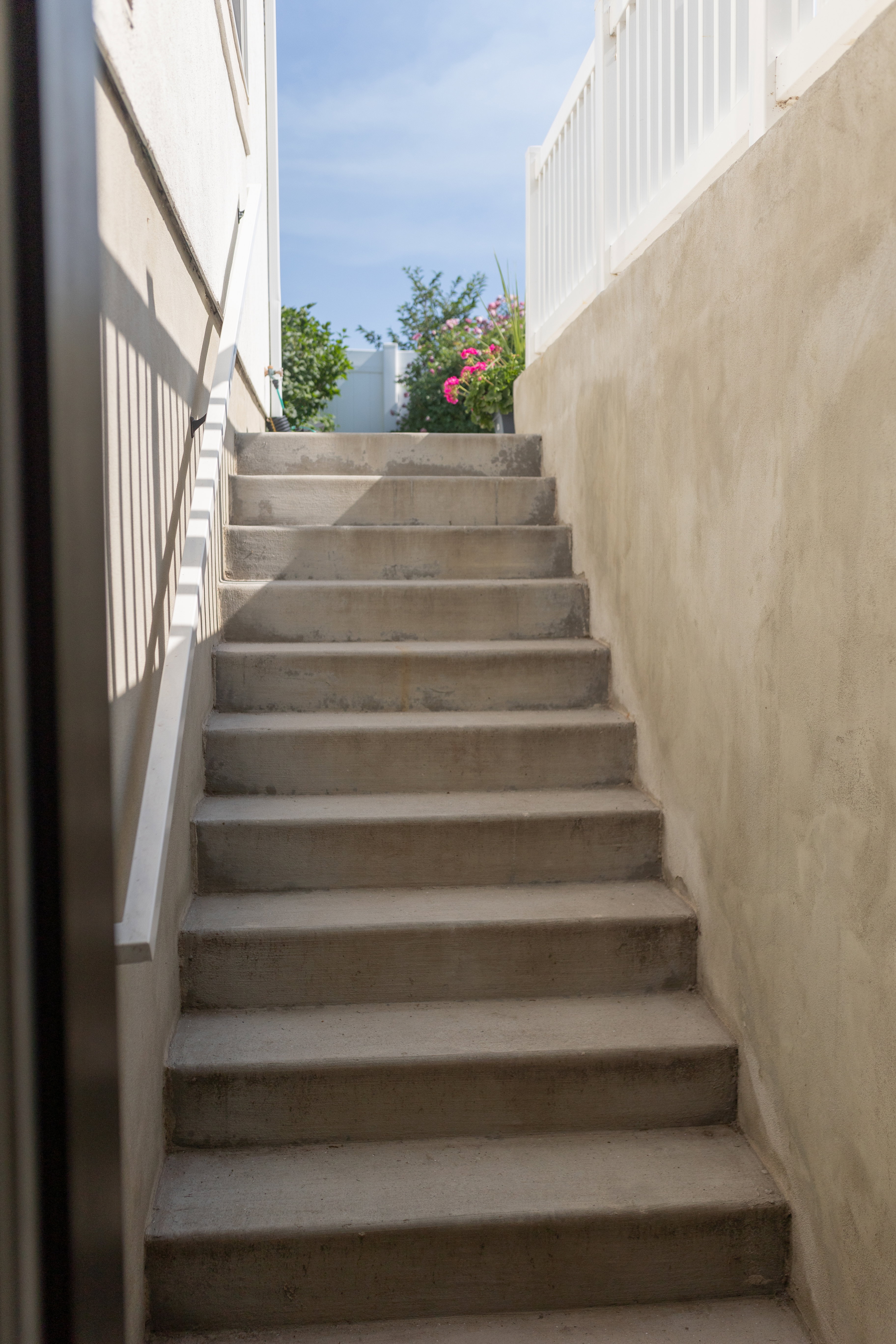 Basement mother-in-law apartment entrance on north east side of home. Concrete staircase with a metal handrail, bordered by a stucco wall and a white picket fence.