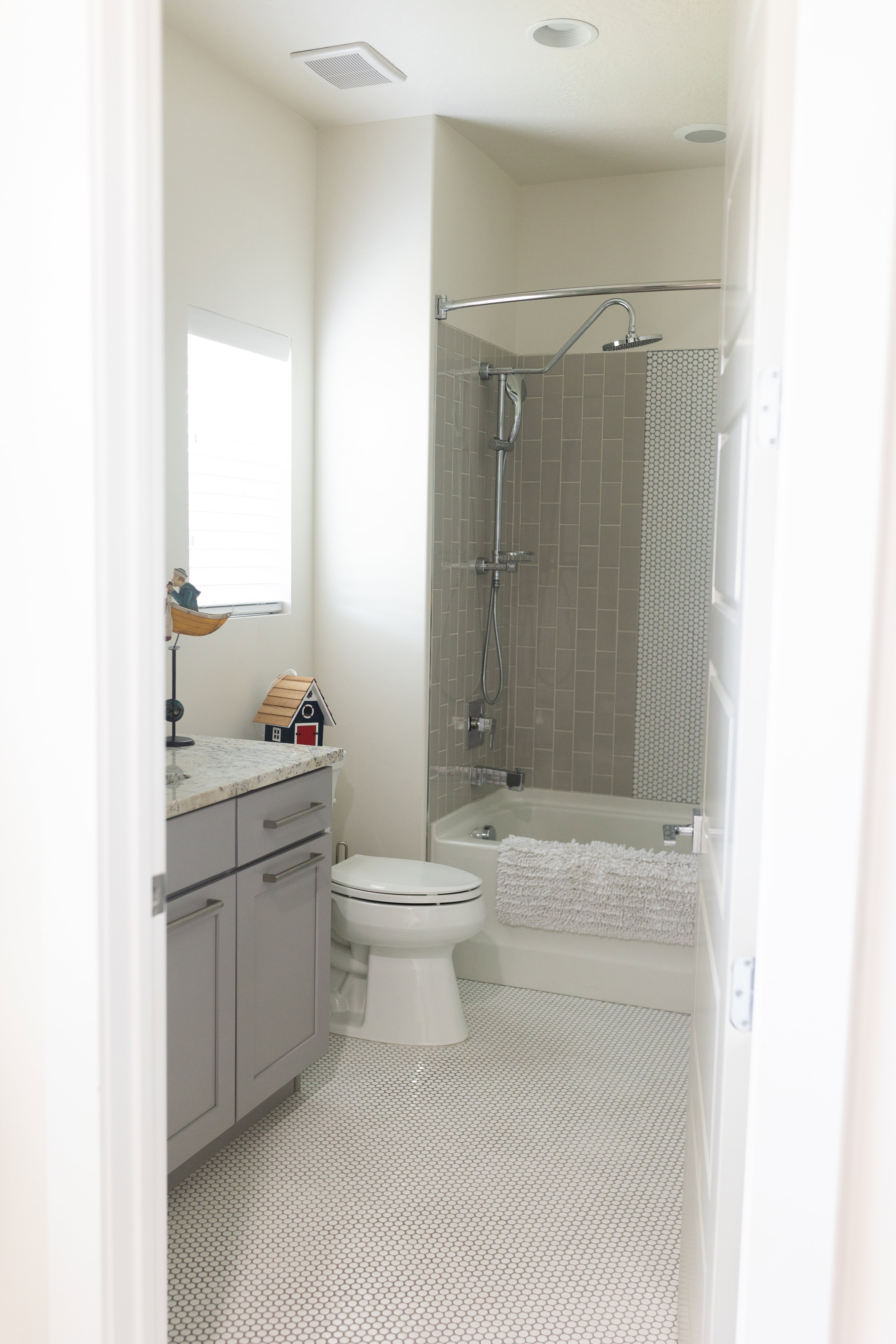 Main floor bathroom featuring white penny tile flooring, a built-in bathtub with subway tile surround, a toilet, and a vanity with a light gray cabinet and solid surface countertop