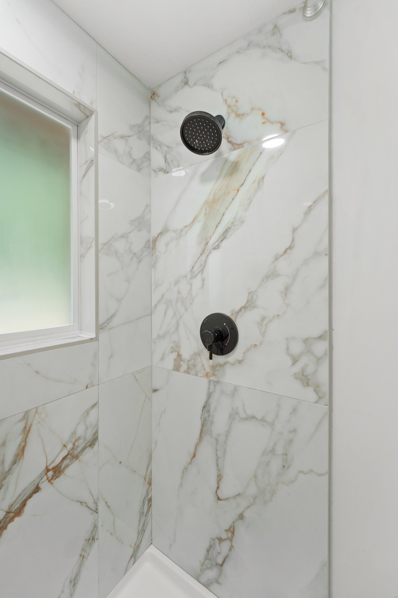 Shower enclosure featuring marble-look tile, matte black shower head, coordinating matte black control handle, and a frosted window with white trim