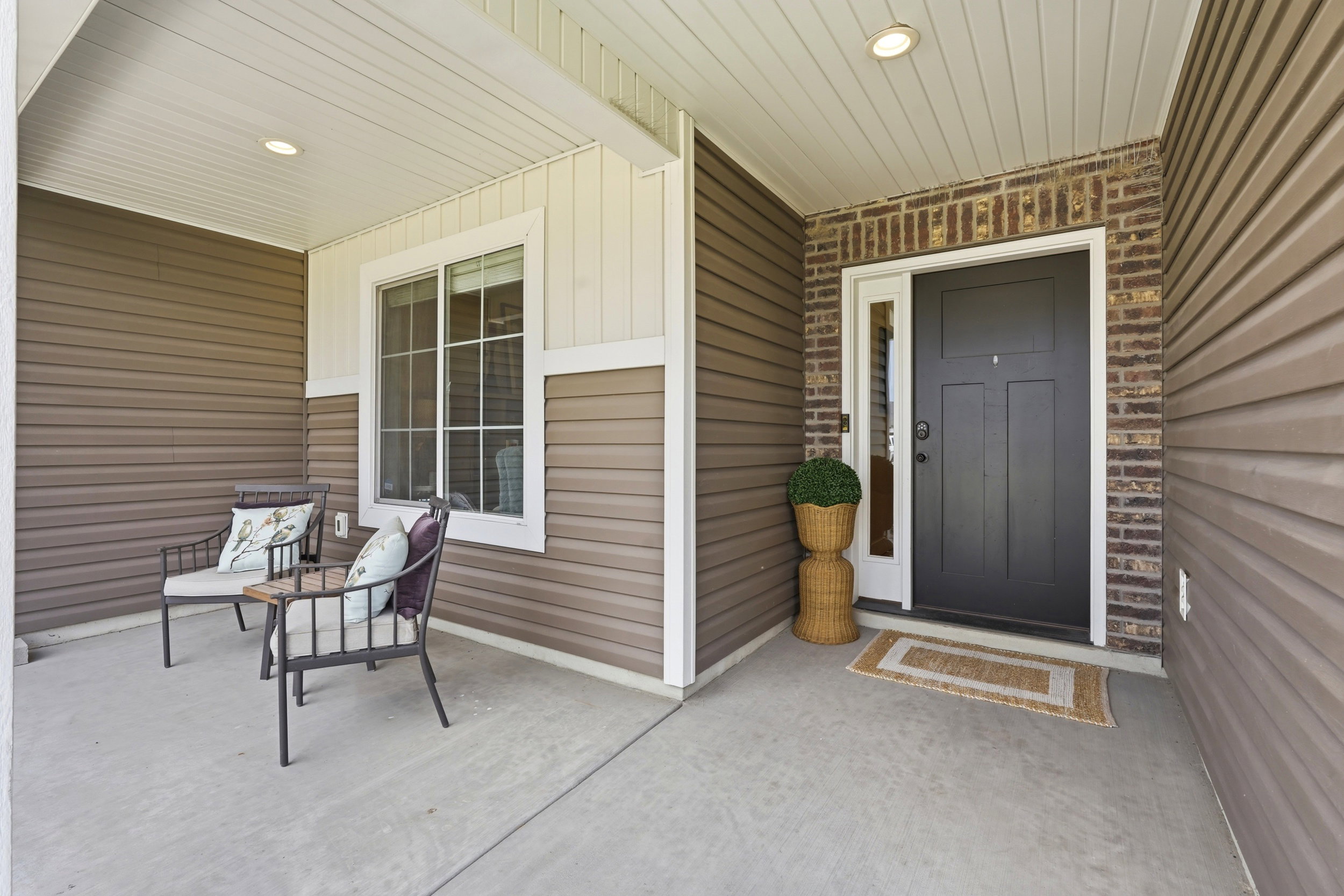 Covered concrete front porch featuring recessed lighting, brown horizontal siding, a dark paneled entry door with sidelight, and brick detailing around the entry