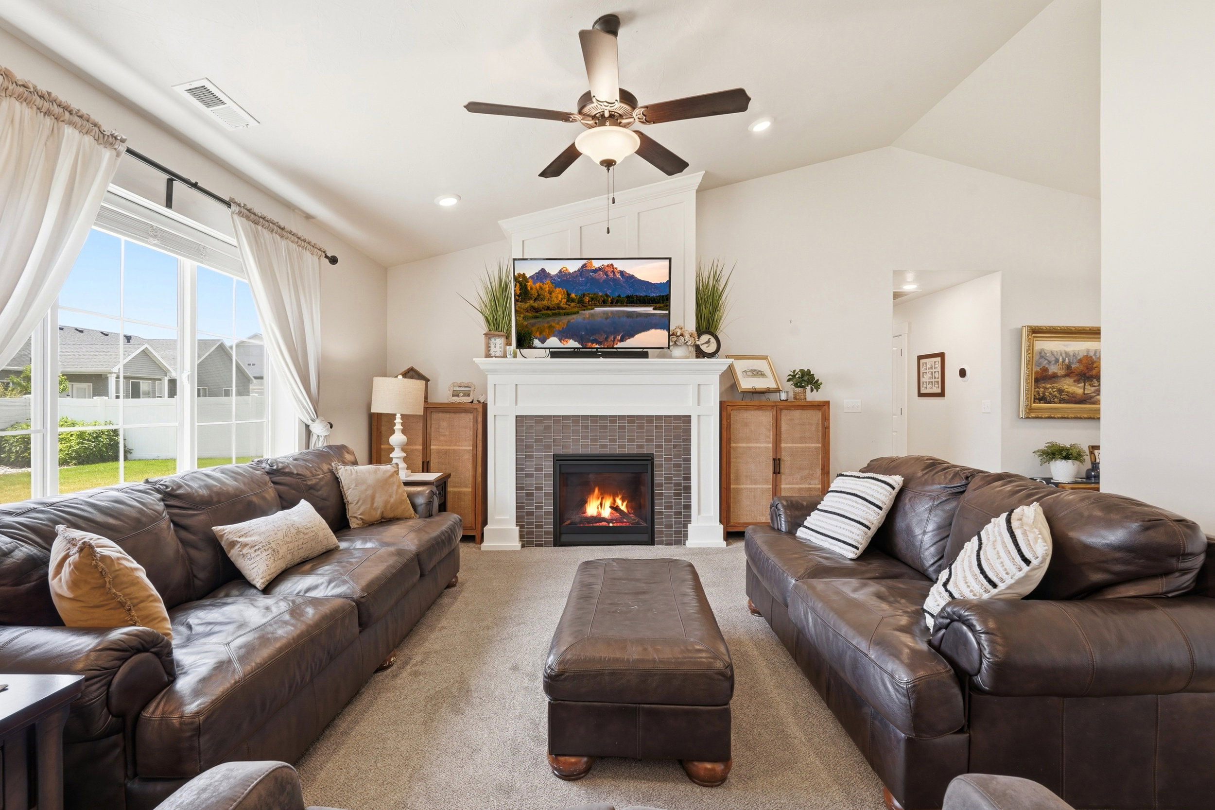 Vaulted ceiling living room featuring a tiled fireplace with a white mantel, ceiling fan, large windows, and neutral carpeting