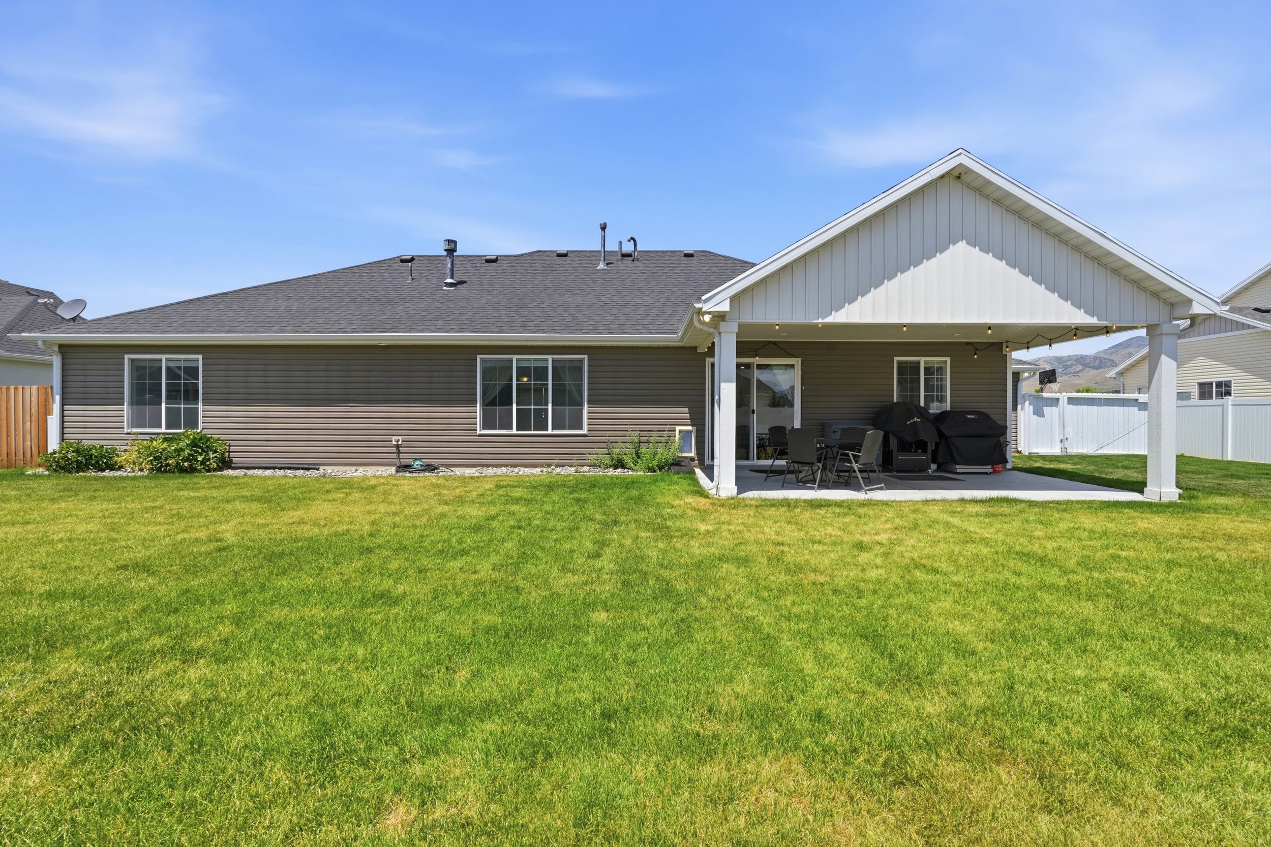 Spacious backyard featuring a covered patio, expansive lawn, white vinyl fencing, and a distant mountain backdrop