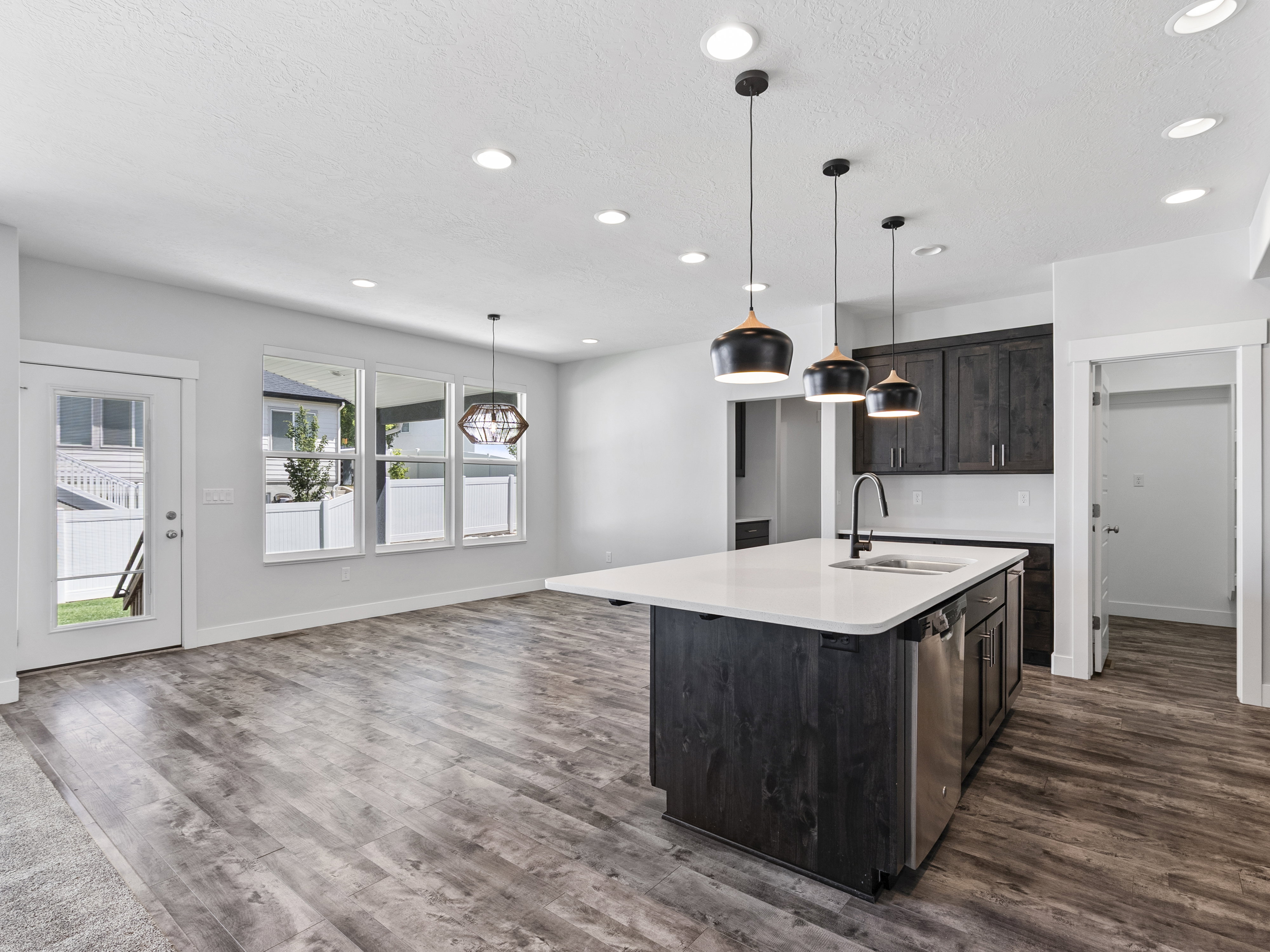 Kitchen island with a white countertop and integrated sink, stainless steel dishwasher, dark wood-finish cabinetry, recessed ceiling lights, and pendant lighting