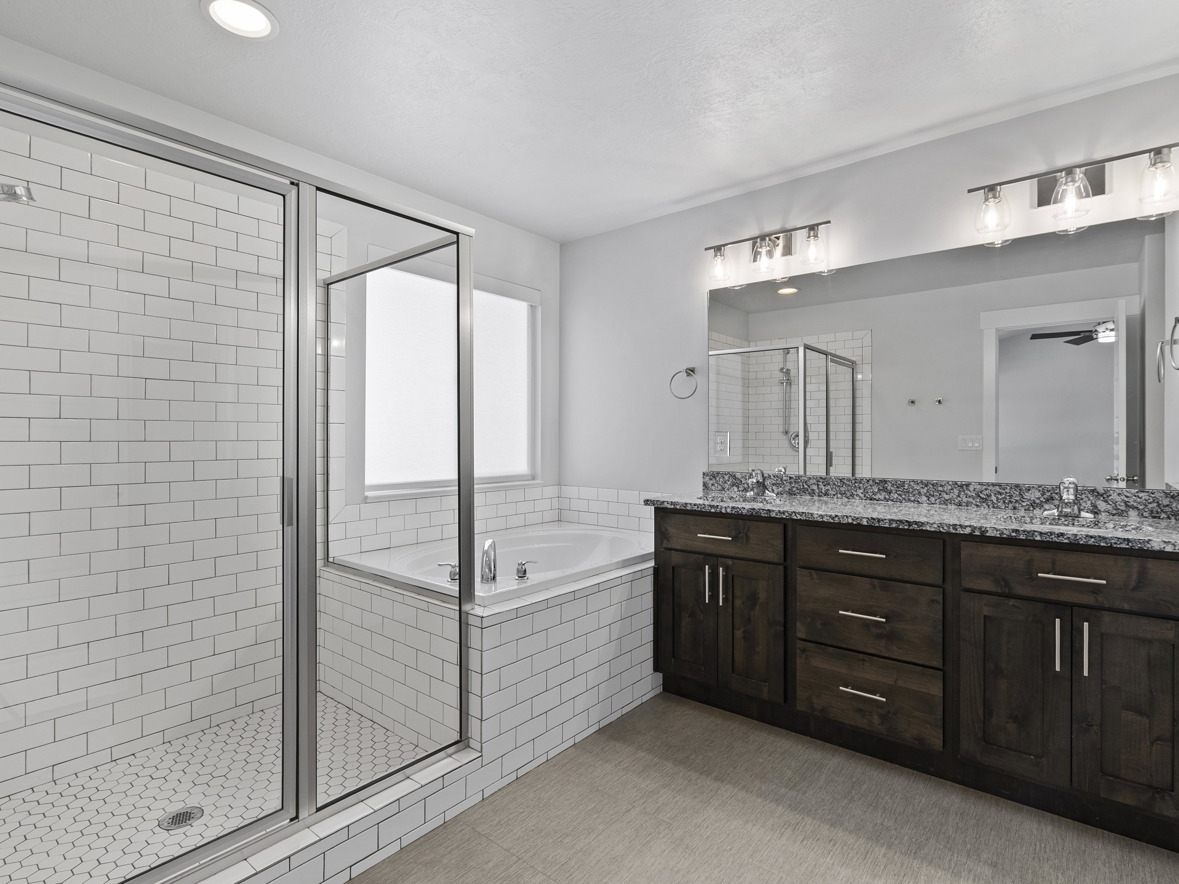 Spacious bathroom featuring a glass-enclosed shower with white subway tile, a built-in tub, dual sink vanity with a stone-finish countertop, dark wood-finish cabinetry, and recessed lighting