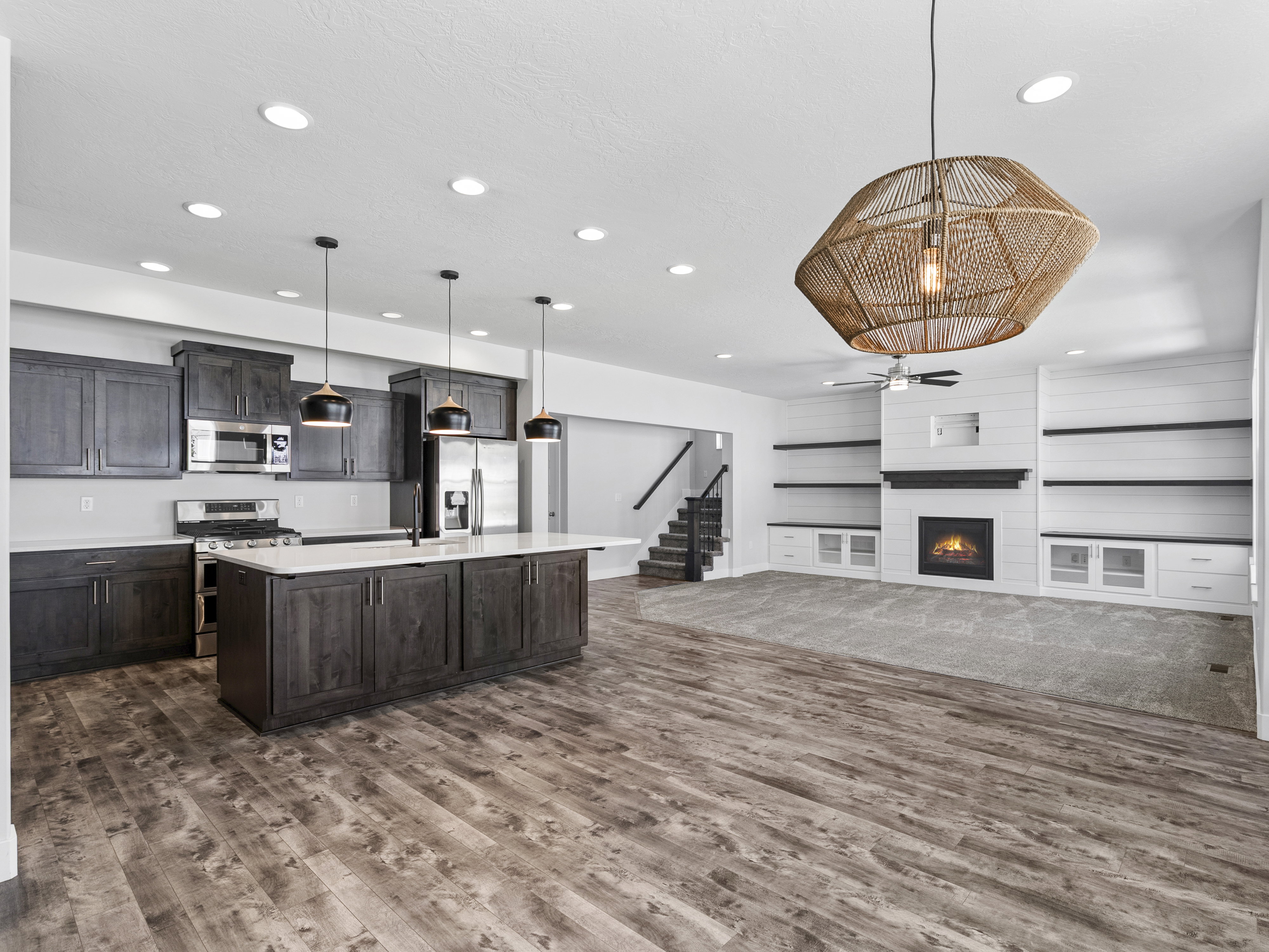 Open-concept living space featuring wood-finish flooring, a kitchen island with quartz countertops, dark wood cabinetry, a built-in fireplace with shiplap detailing, and integrated shelving with lower cabinetry