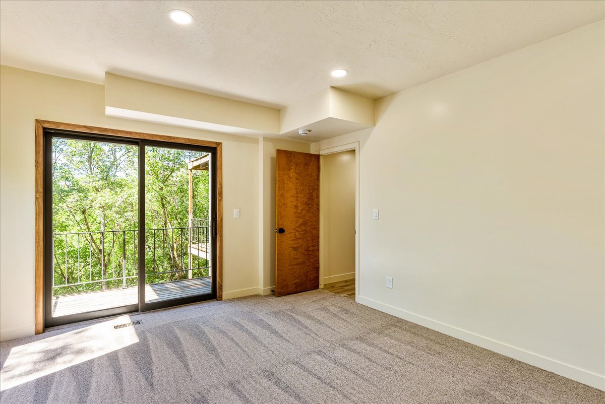 Carpeted room with a sliding glass door leading to a balcony, featuring a dark metal railing and surrounded by mature trees
