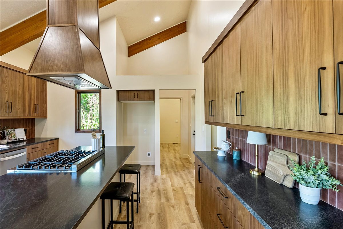 Spacious kitchen featuring a large island with a gas range, wood-finish cabinetry, dark countertops, a unique tile backsplash, and vaulted ceilings with exposed beams