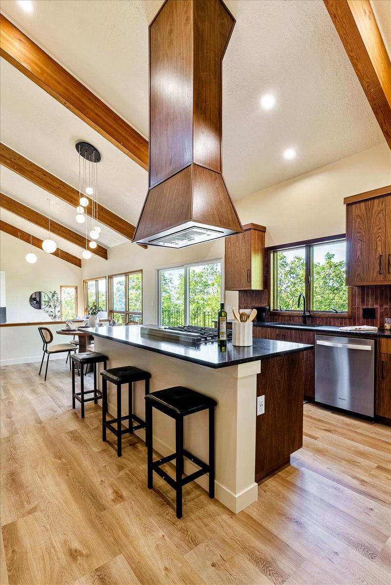 Open-concept kitchen featuring vaulted ceilings with exposed wood beams, a large island with dark countertops, wood-finish cabinetry, and stainless steel appliances