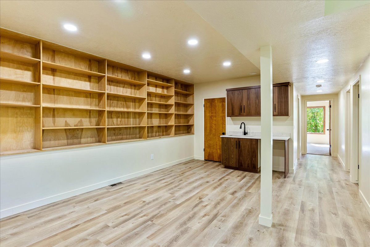 Spacious room featuring extensive built-in wooden shelving, wood-finish flooring, recessed lighting, and a wet bar with dark wood cabinetry, a white countertop, and a matte black faucet