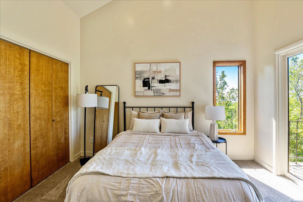 Vaulted ceiling room featuring light-toned walls, wood-finish bi-fold closet doors, a tall window with wood trim, and a glass-paneled door opening to an exterior space