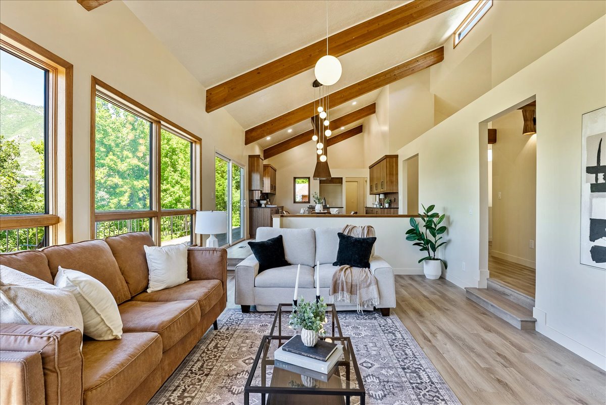 Light-filled living area featuring vaulted ceilings with exposed wood beams, expansive windows, wood-finish flooring, and a contemporary chandelier