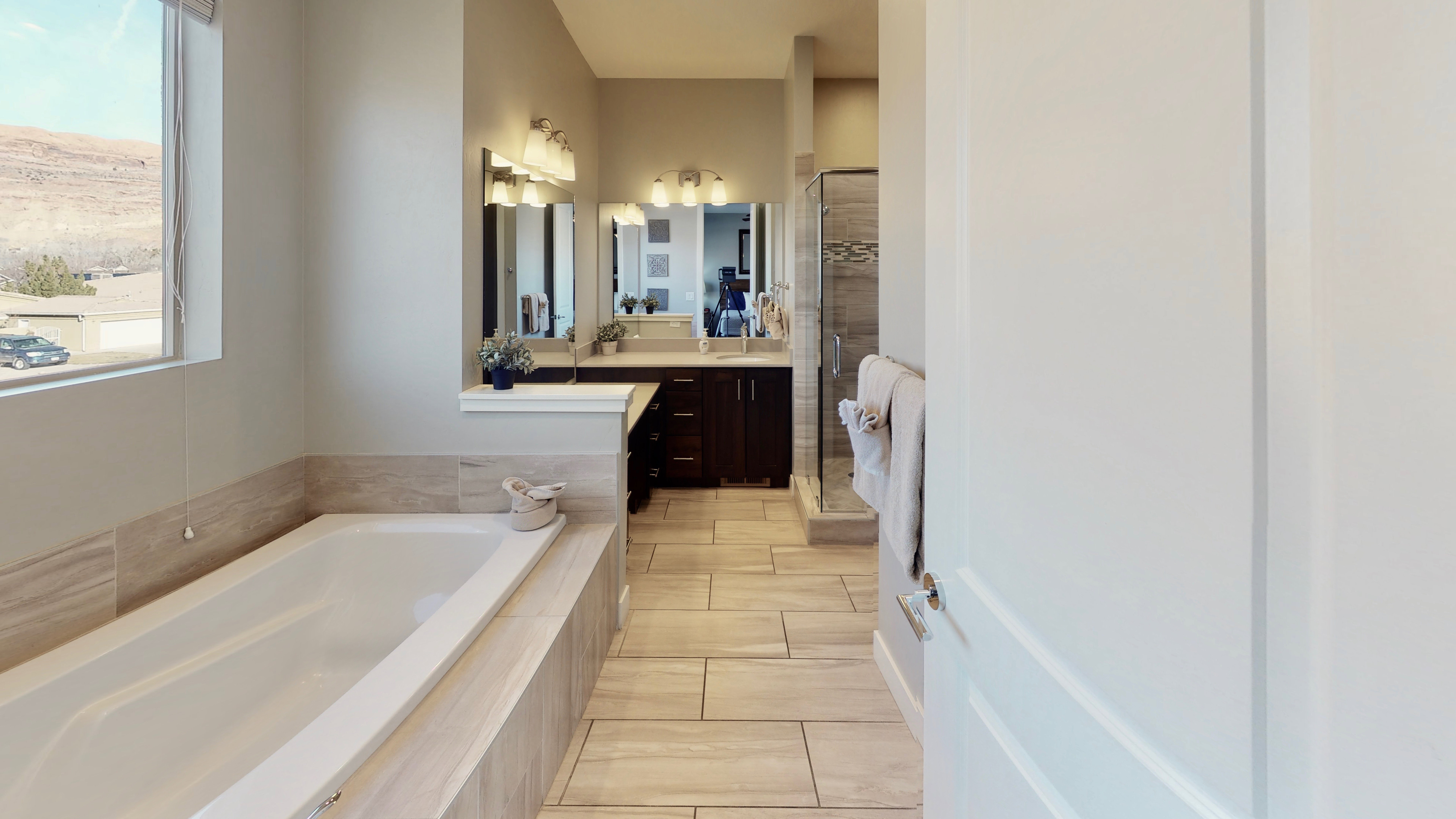 Soaking tub with tile surround, dual vanity with dark wood cabinetry, framed mirrors, and a glass-enclosed shower