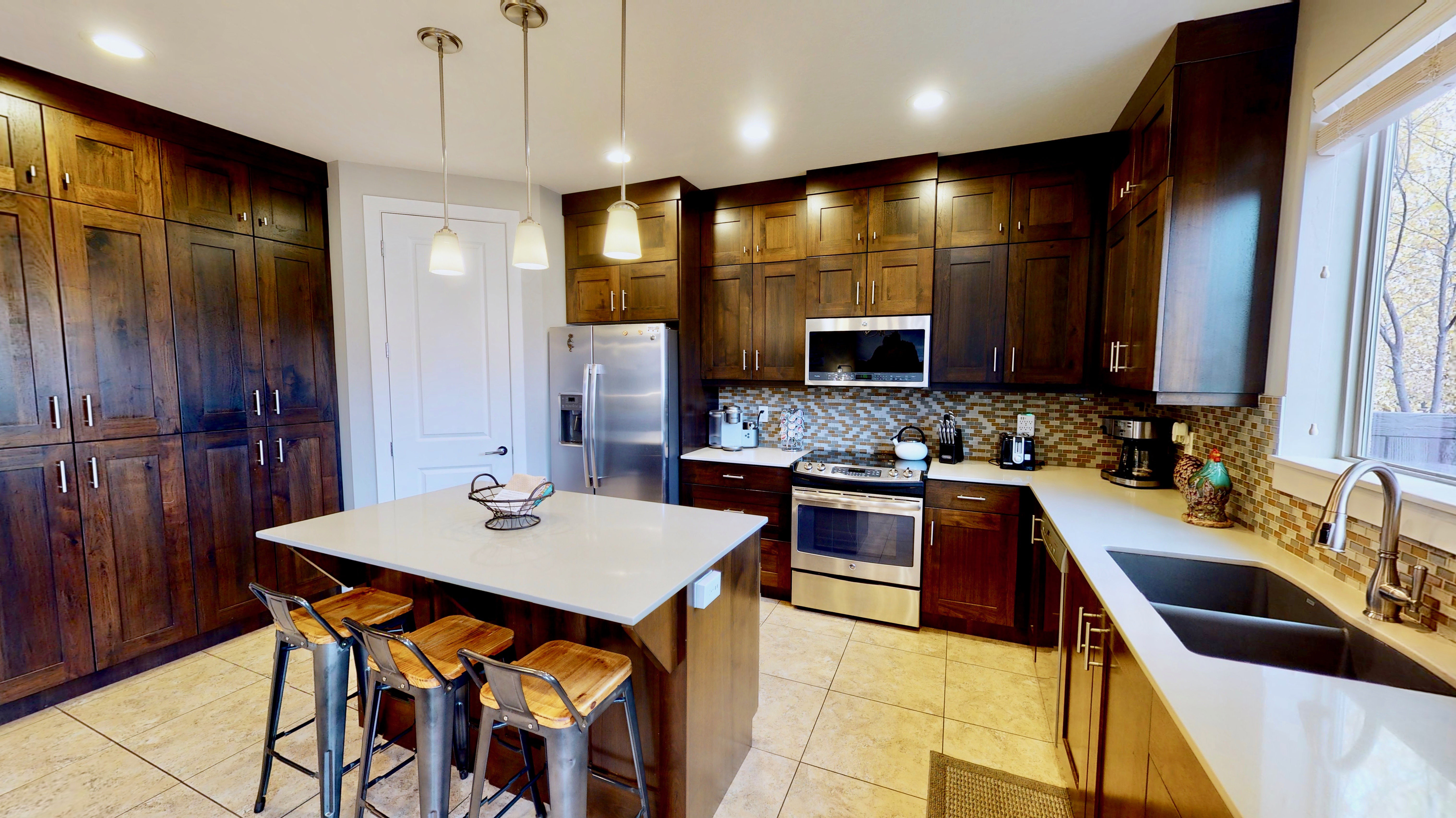 Modern kitchen featuring dark wood cabinetry, a central island with countertop seating, stainless steel appliances, a mosaic tile backsplash, and a double basin sink with a gooseneck faucet