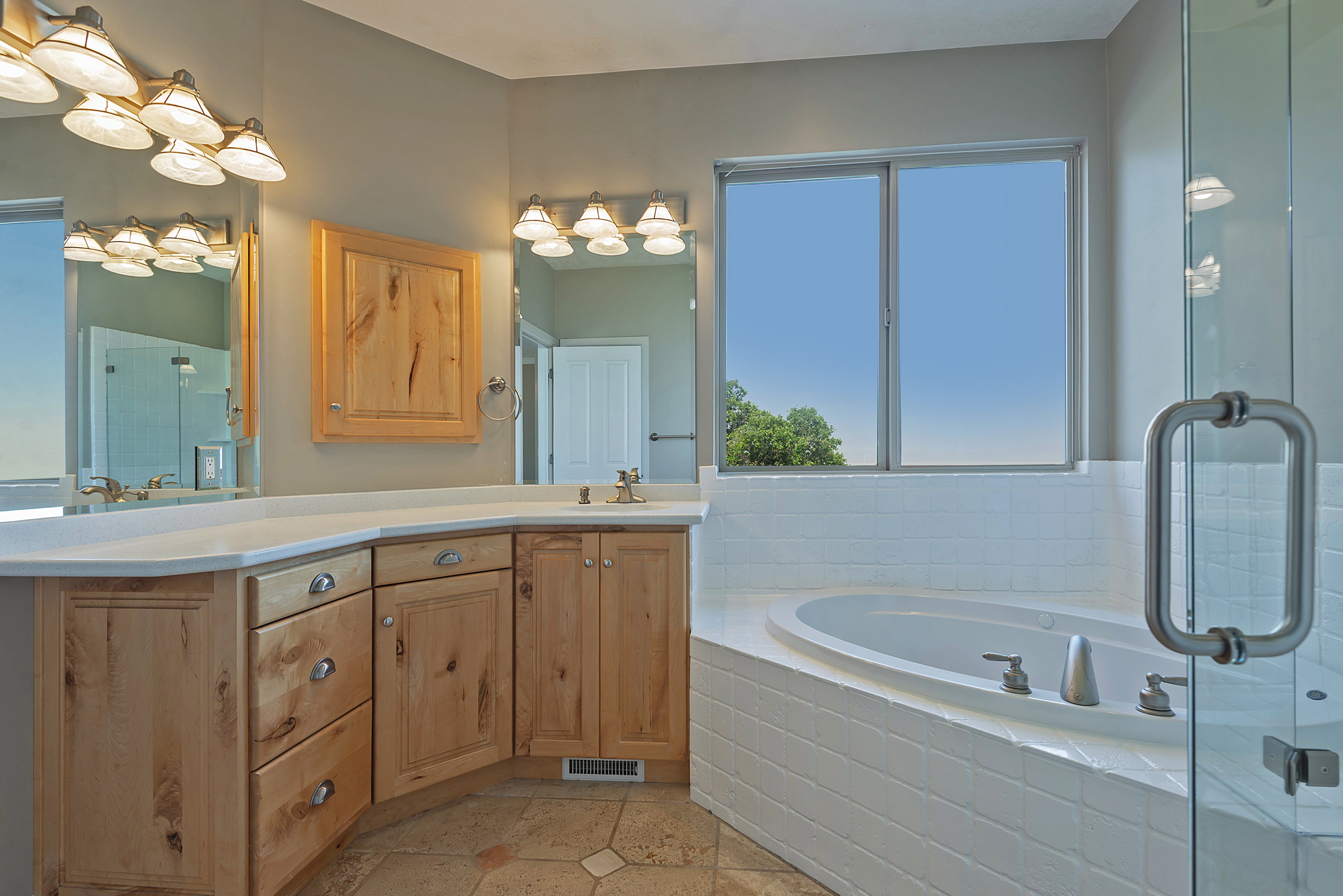 Bathroom featuring a built-in jetted tub with white tile surround, a glass-enclosed shower, a double vanity with wood cabinetry, and a large window