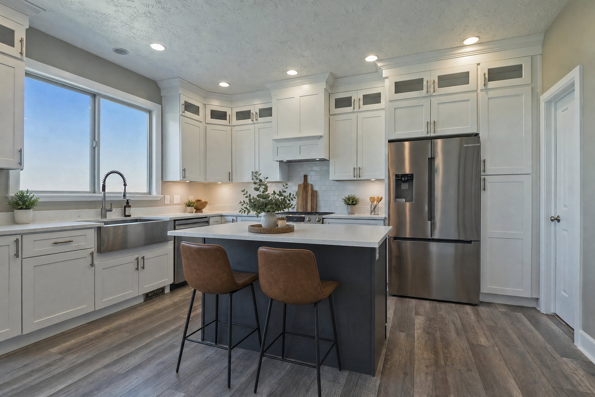 Modern kitchen featuring white cabinetry, a large window above a stainless steel farmhouse sink, a central island with a light-toned countertop, stainless steel appliances, and wood-finish flooring